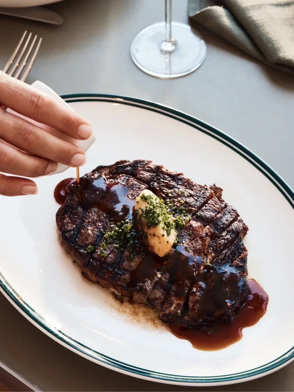 Close-up of grilled steak topped with herb butter and rich sauce, being finished tableside on a white serving plate.