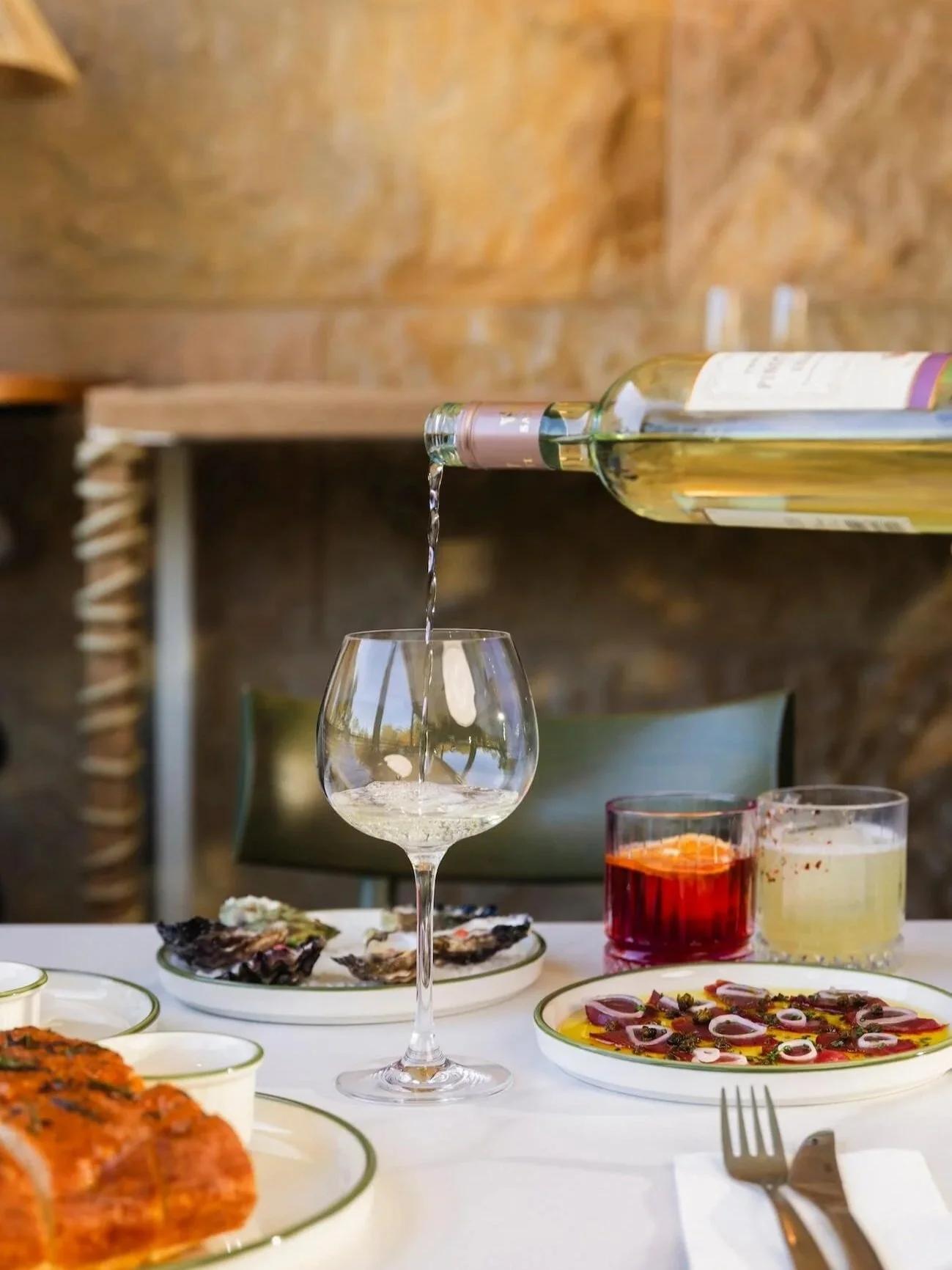 white wine being poured into a crystal glass at a white-clothed dining table set with green-rimmed plates, oysters, a beef carpaccio, focaccia, a red Negroni and a yellow cocktail, against a warm stone wall.