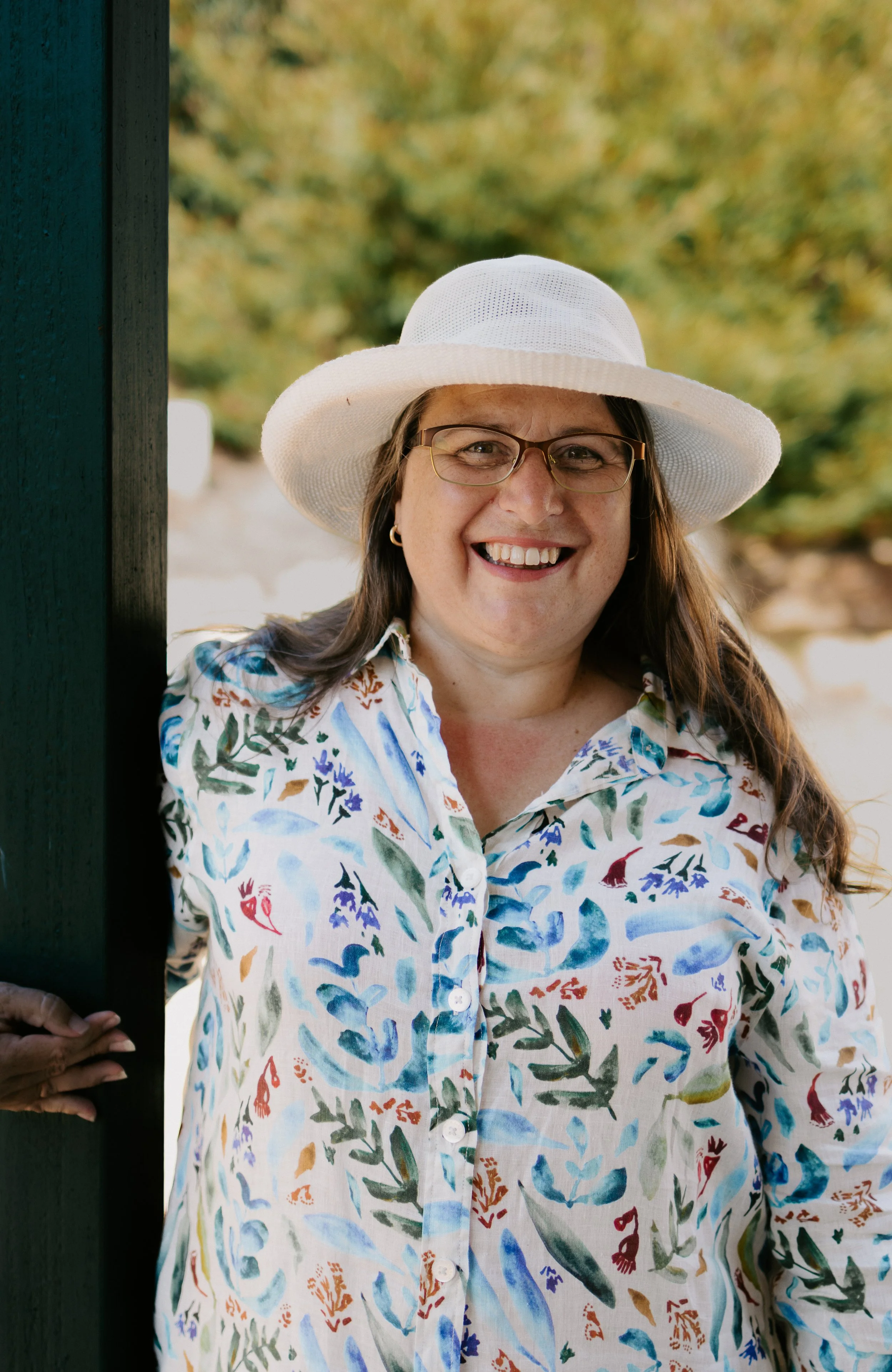 A smiling woman wearing glasses, a white hat, and a colorful floral shirt standing outdoors near a green painted structure with trees in the background.