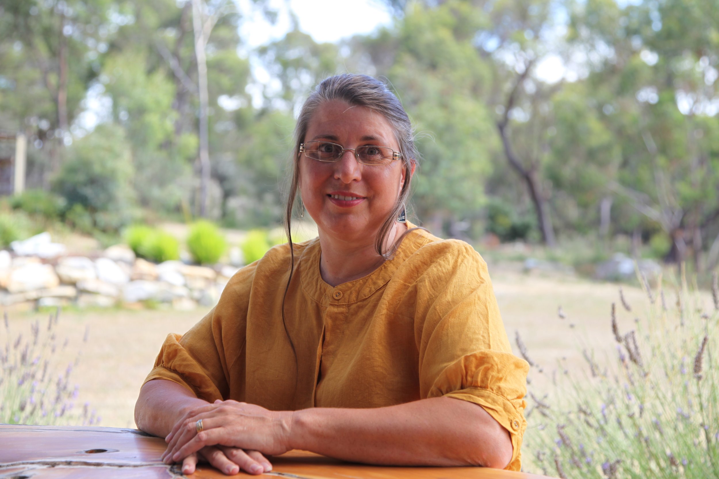 Amanda Erskine  money energy intuitivesmiling with glasses and earrings, wearing a yellow blouse, is sitting outdoors at a wooden table, with her arms crossed on the table. The background features a blurred natural setting with trees