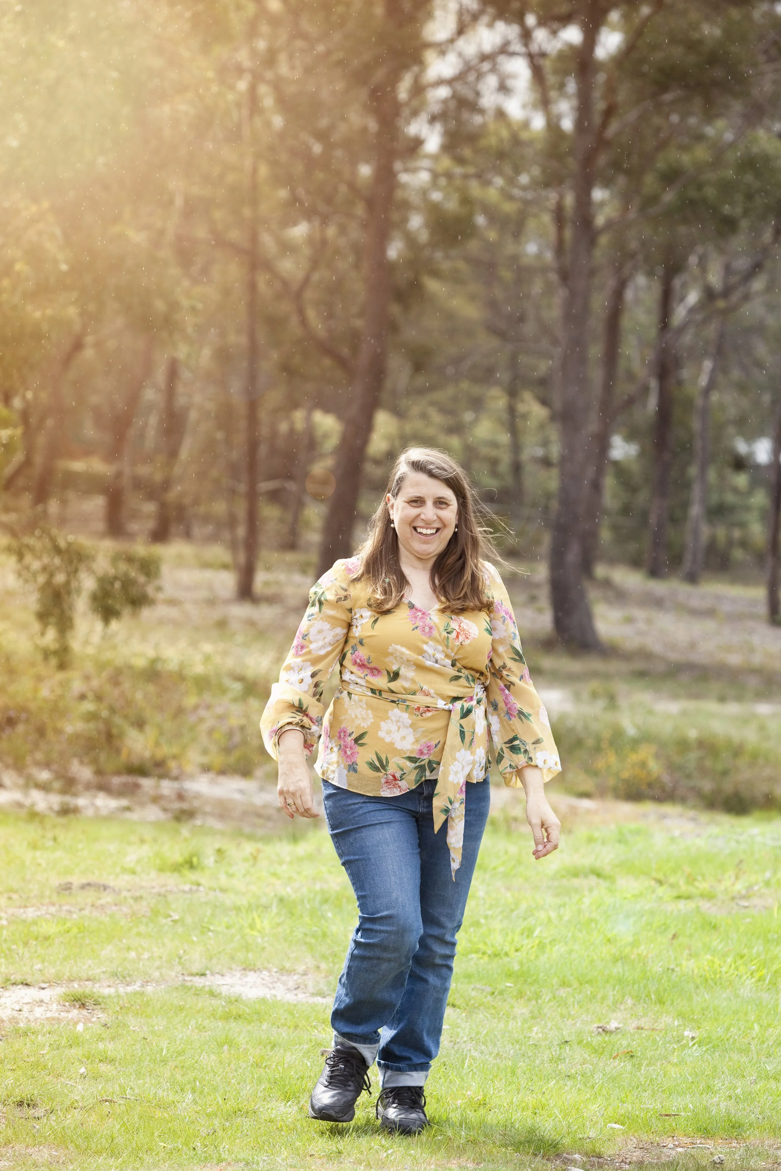 Amanda Erskine smiling and walking in a rain shower grass in a park with trees in the background, wearing a yellow floral blouse and blue jeans - Money Energy healing practitioner