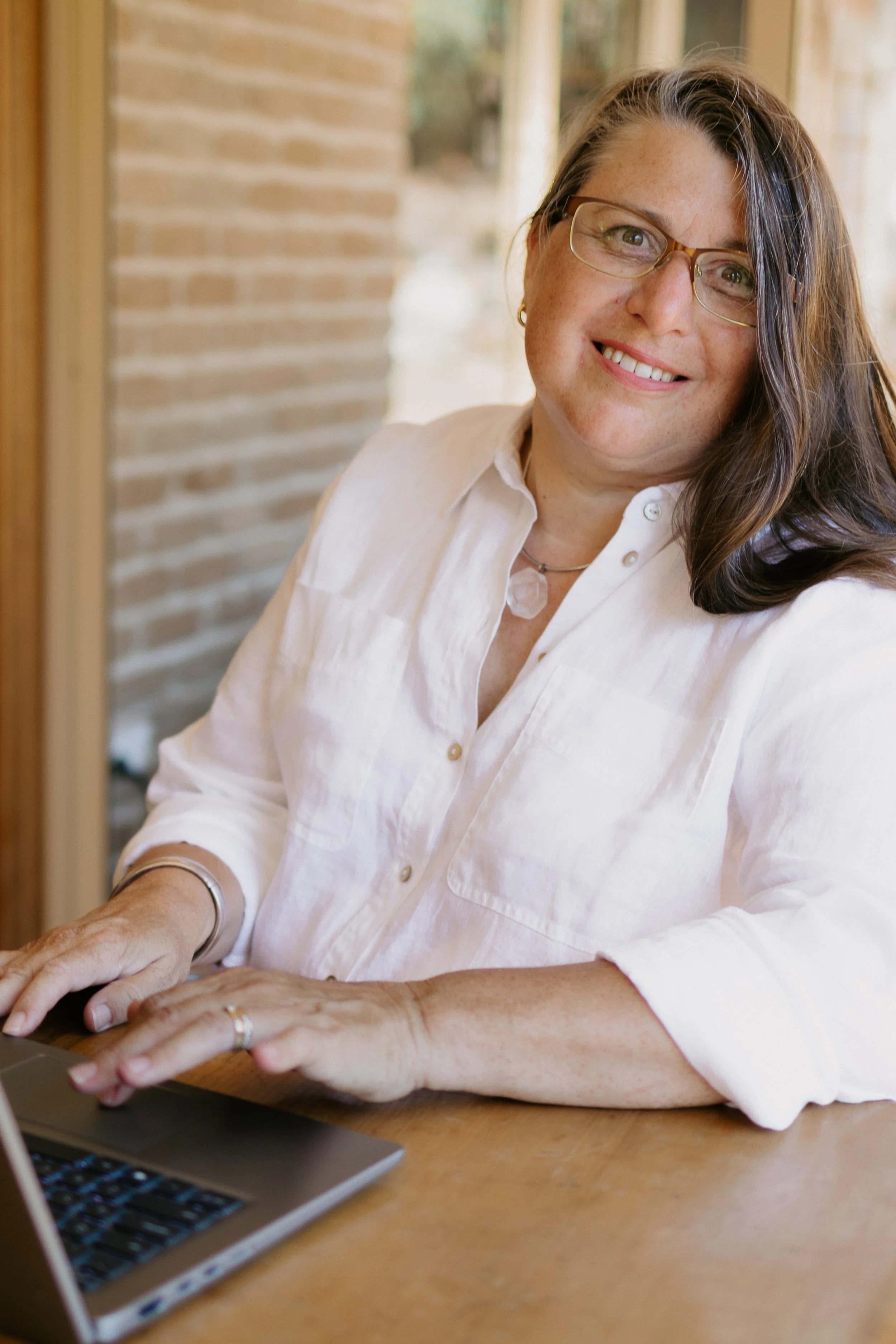 A woman with glasses and a white shirt sitting at a wooden table, smiling, working on a laptop.