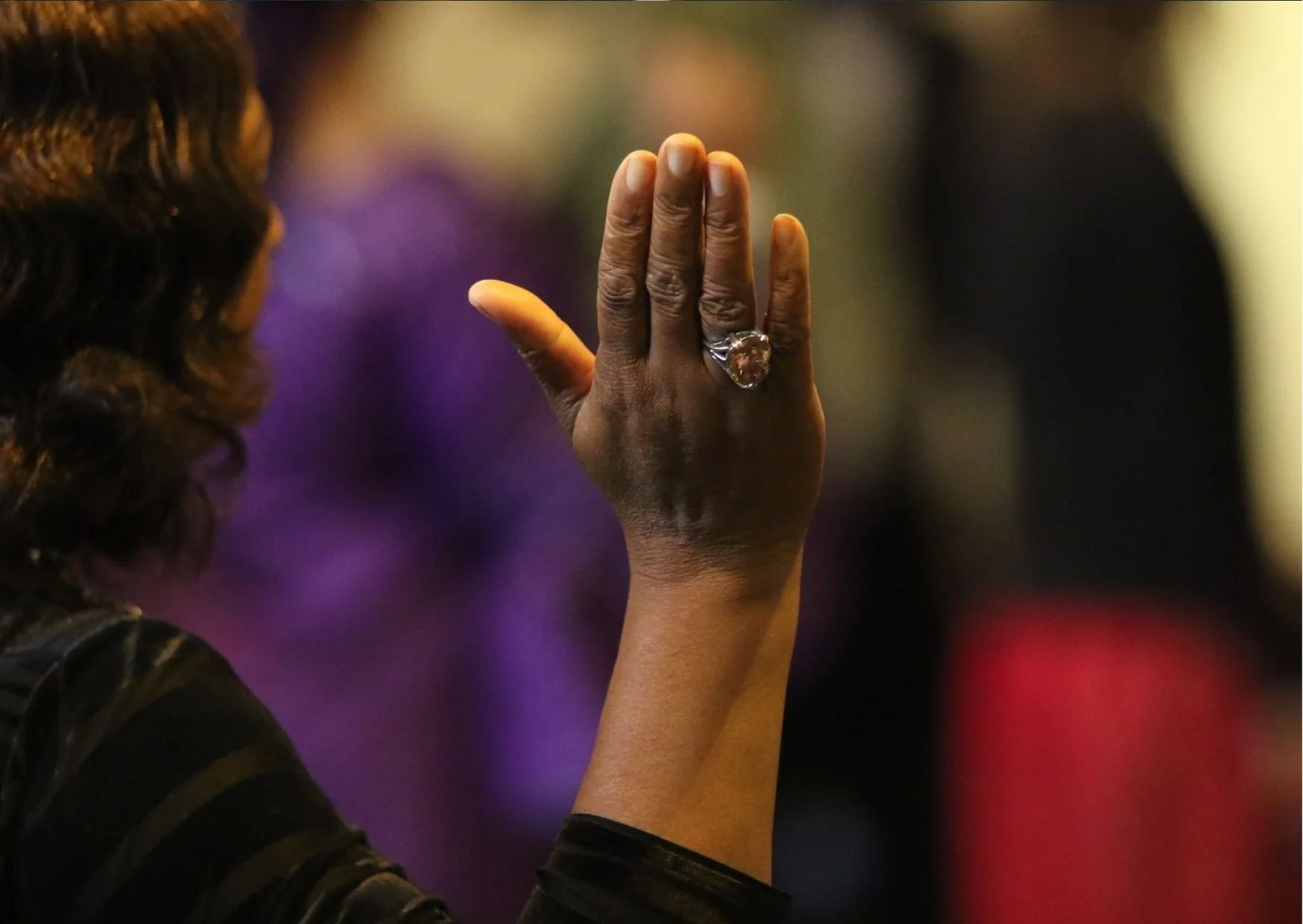 A woman with dark skin and dark hair is shown from the side, raising her hand with her fingers together. She is wearing a large heart-shaped ring. The background is blurred with purple, black, and red colors.