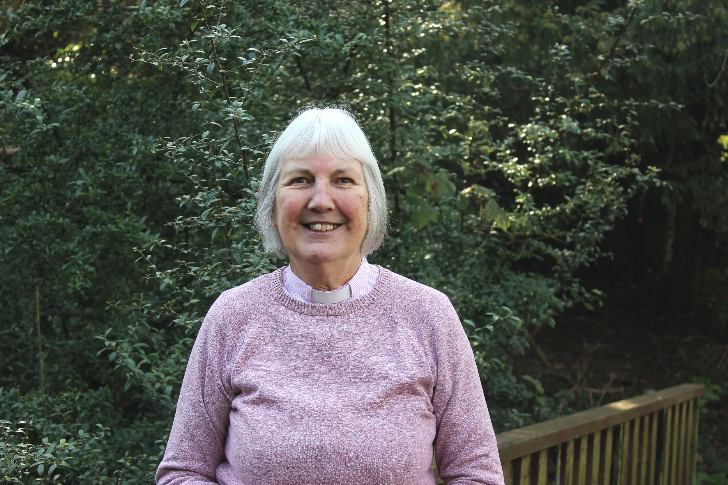 A smiling elderly woman with short white hair, wearing a teal clerical shirt with a white collar, holding a sign that reads 'Elaine Weaver Associate Vicar' in front of a stone building.