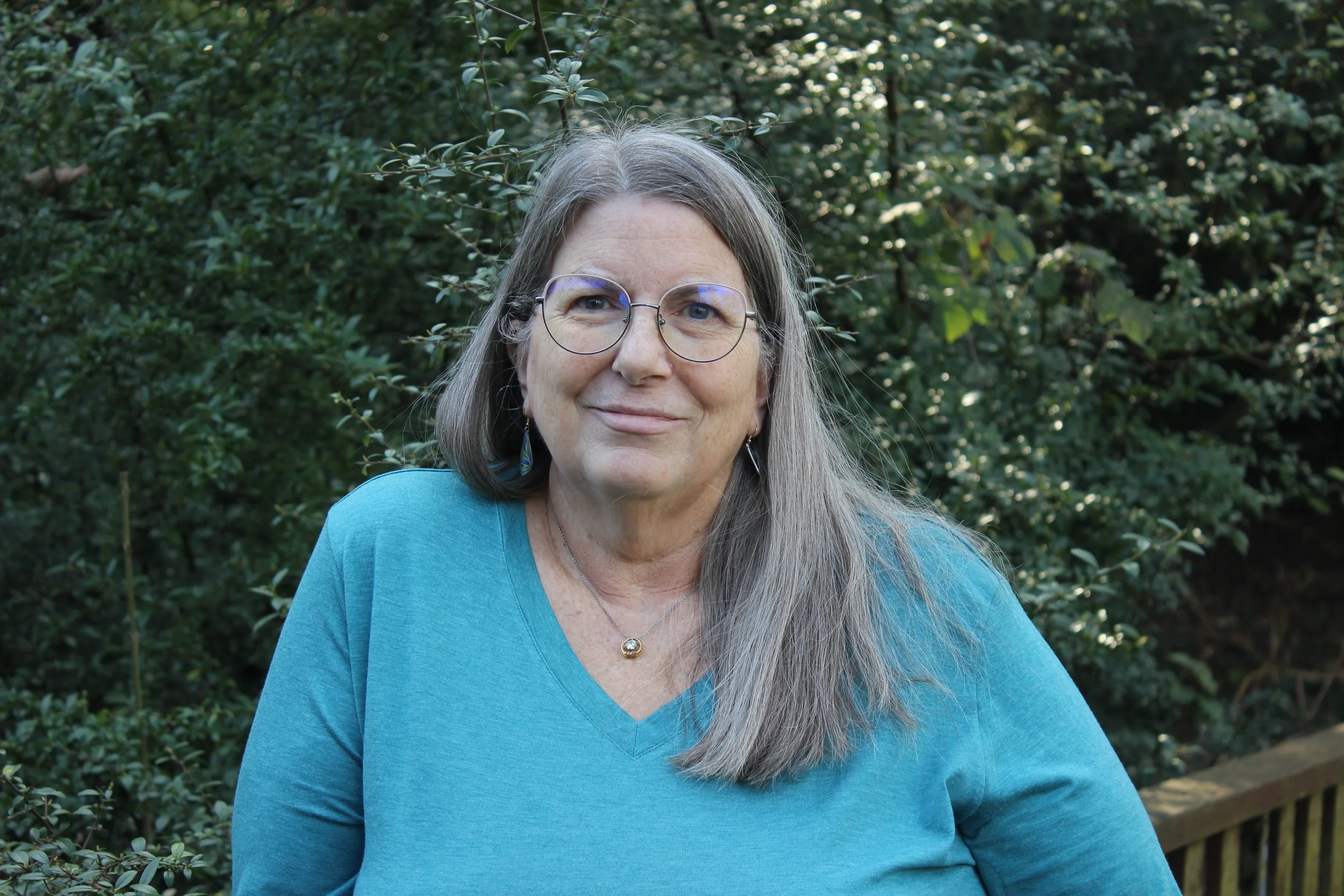 A smiling woman with glasses and long gray hair holding a sign that reads 'Barbara Kuekes-Patel, Administrator' in front of a garden with green plants and red flowers.