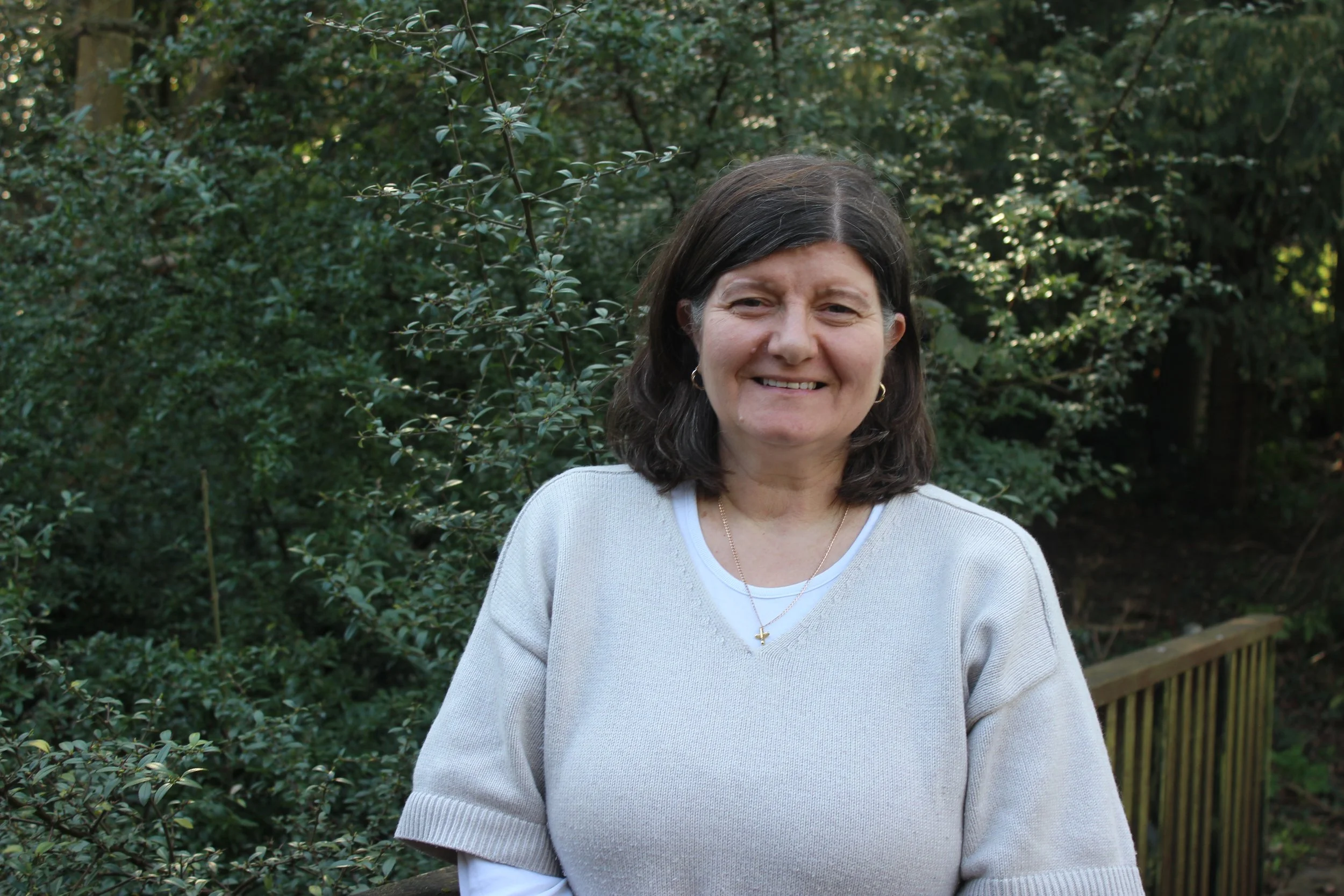 A woman smiling and holding a sign that reads 'Carol Sormaz Children & Families Worker' outdoors with green foliage in the background.