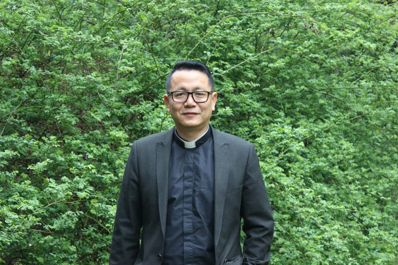A man wearing glasses, a clerical collar, and a denim jacket holding a white sign with the text 'Kailean Khongsai Curate' in an outdoor setting with trees and fallen leaves.