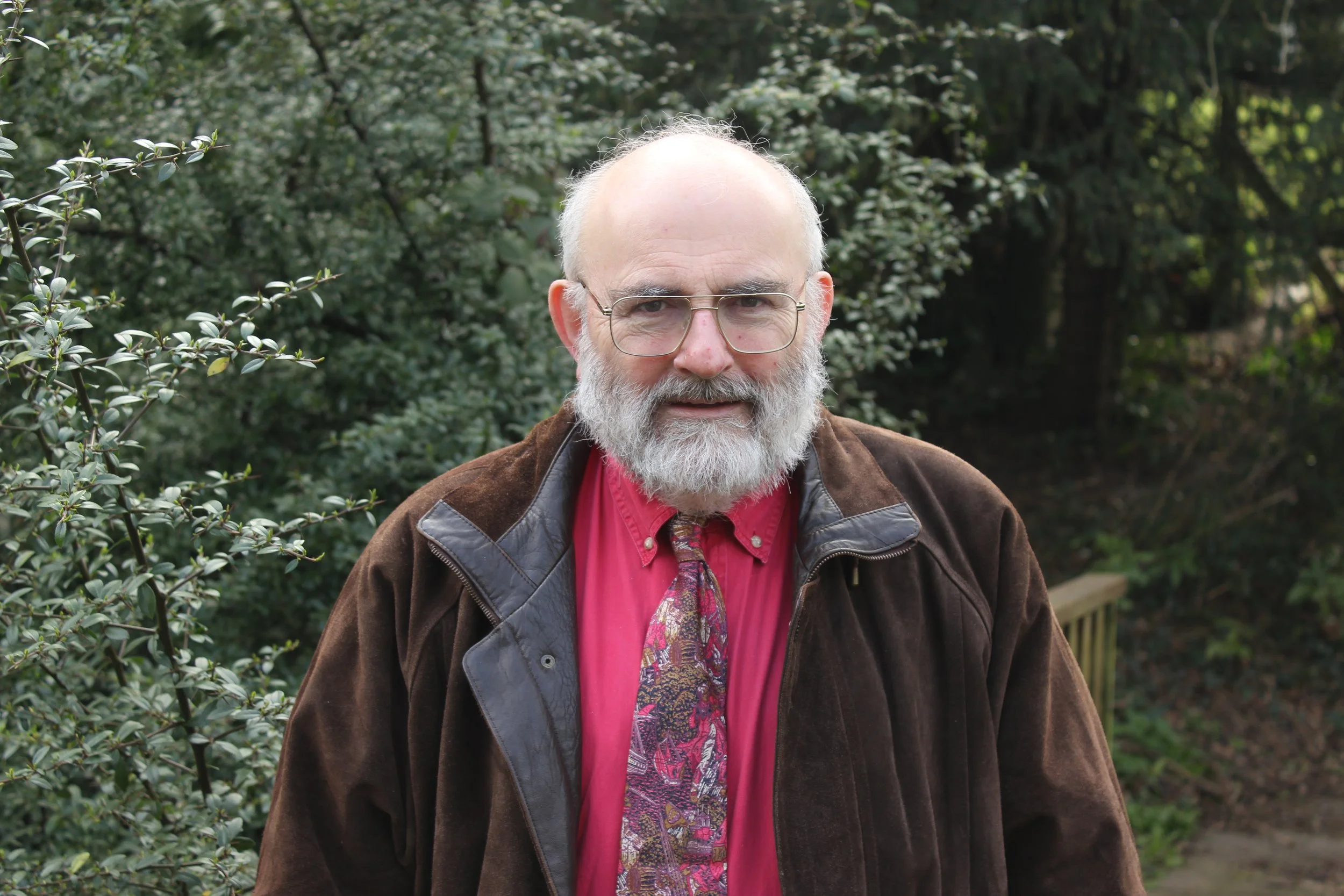 Man with glasses and beard holding a sign that reads 'Clive Scowen Licensed Lay Minister' inside a church or hall with a window showing clothes hanging outside.
