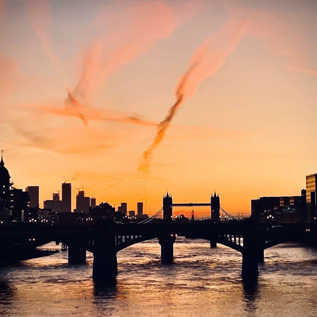 Sunset over the London skyline with the Tower Bridge, city buildings, and contrails in the sky.