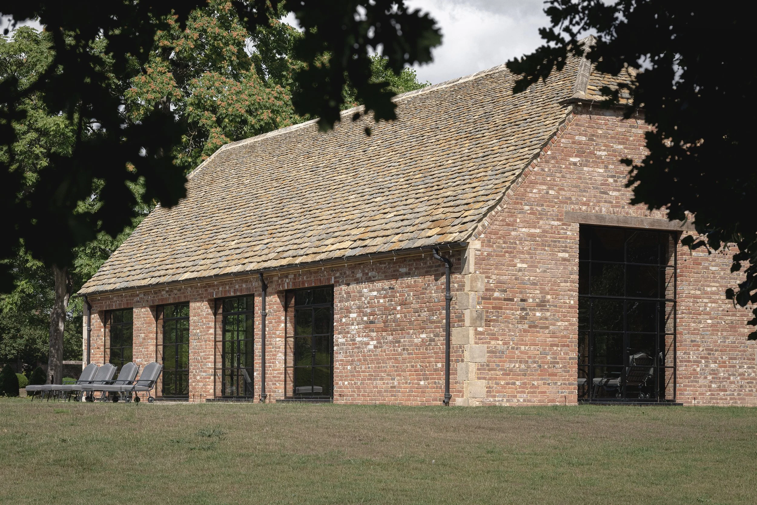 A brick building with large black window frames, a tiled gabled roof, and a small porch. There are trees and chairs outside the building.