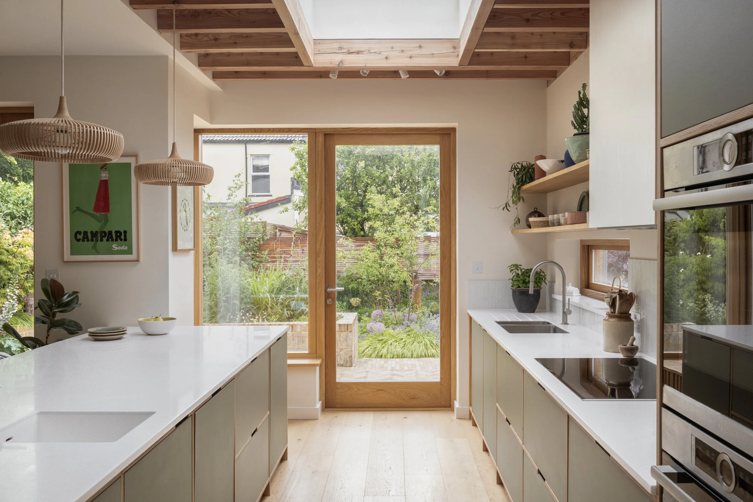 Modern kitchen with white countertops, light wood cabinetry, and large glass door leading to a garden.