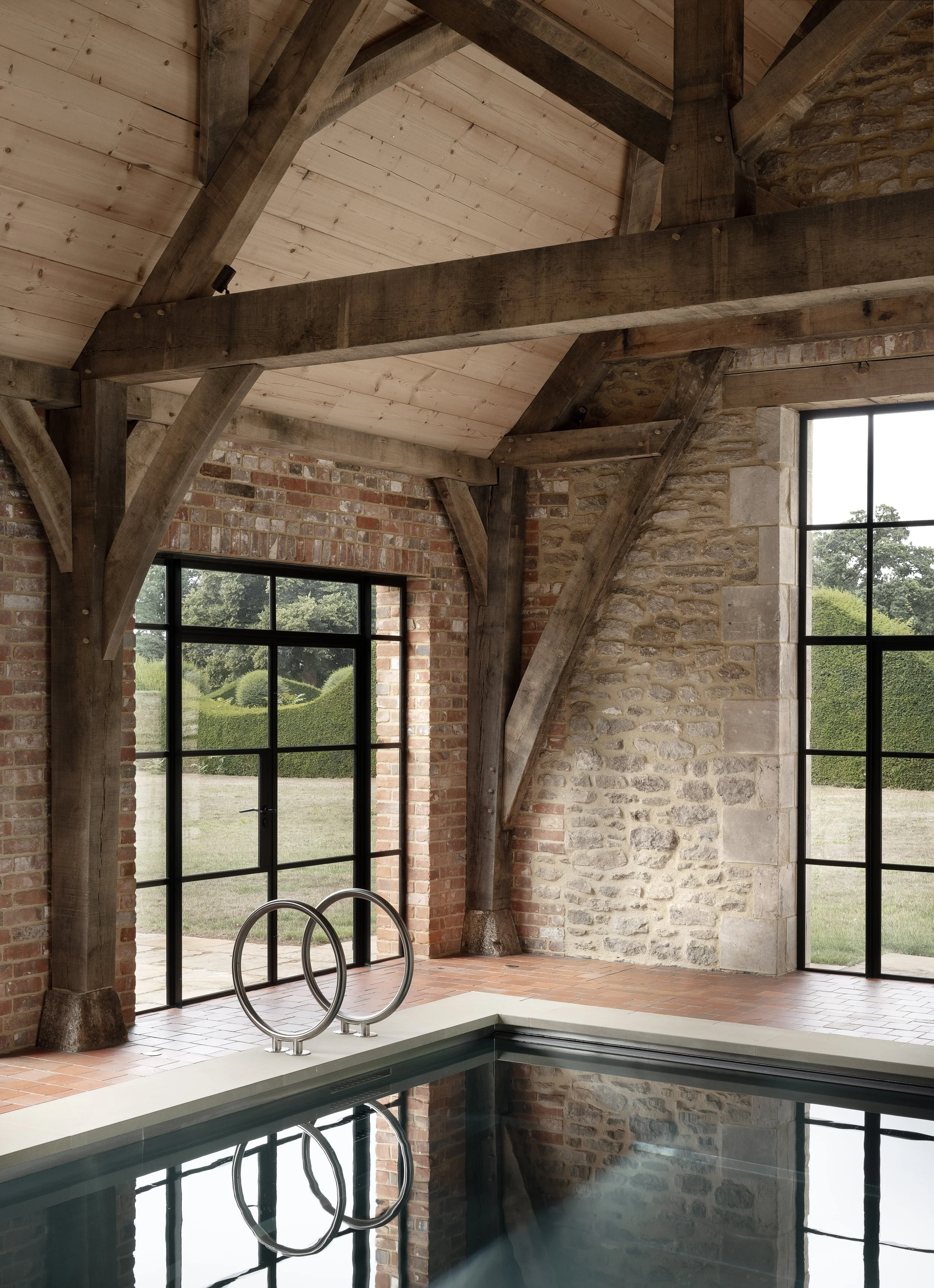 Indoor pool area with large windows, brick and stone walls, wooden beams and ceiling, and metal pool ladder.