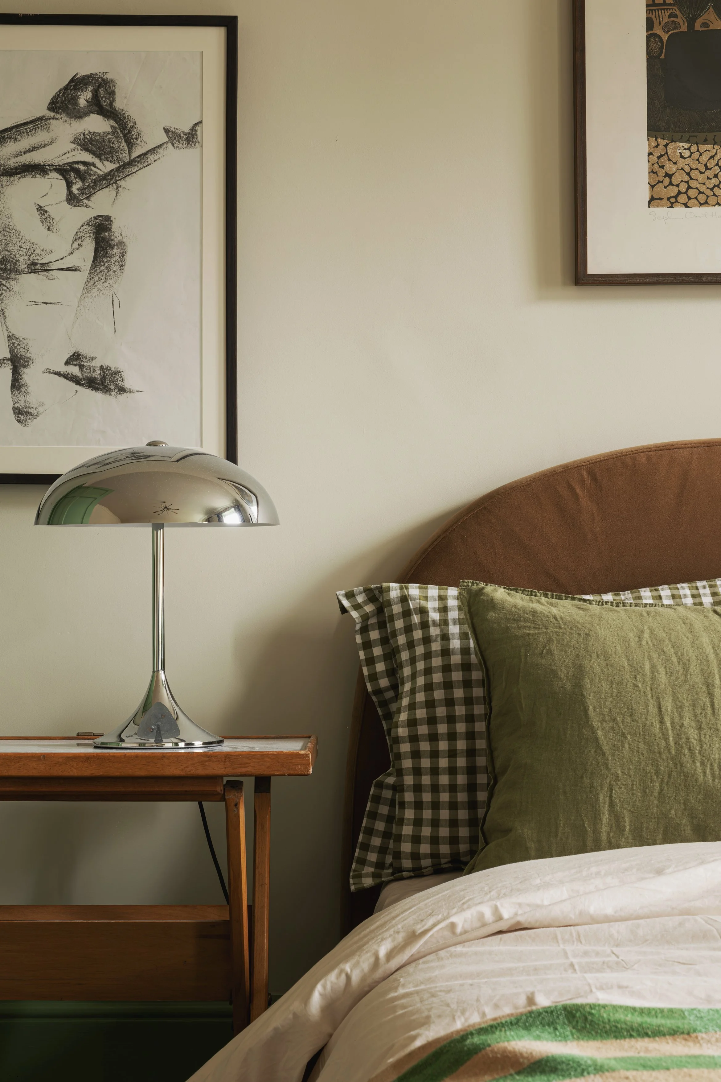 A bedroom corner featuring a wooden side table with a chrome lamp, a bed with green and checkered pillows, and framed artwork on a light-colored wall.