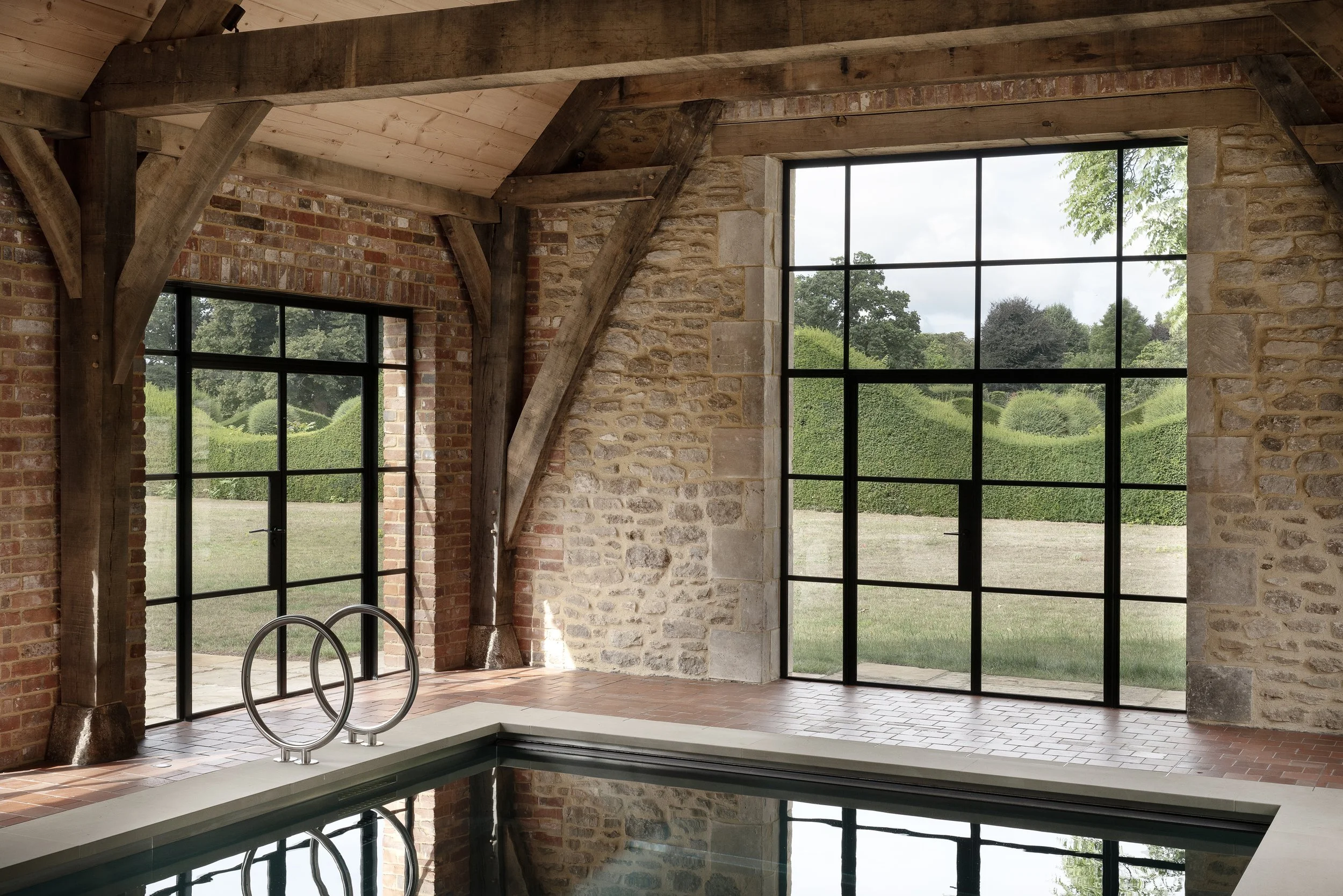 Indoor pool area with large floor-to-ceiling windows showing greenery outside, brick and stone walls, and wooden beams on the ceiling.