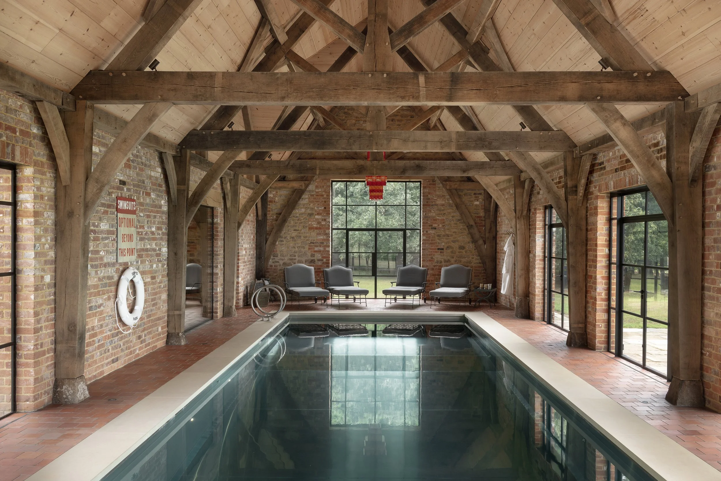 Indoor swimming pool area with brick walls, wooden beams, and large windows showing outdoor greenery. Six lounge chairs are positioned on the far side, and a yellow and red life preserver and a sign are mounted on the left wall.