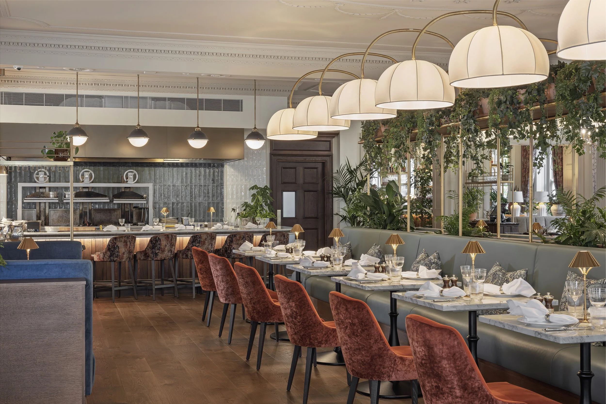 Elegant restaurant interior with white and gold hanging lamps, green plants, and neatly set dining tables with white napkins and gold lamps.
