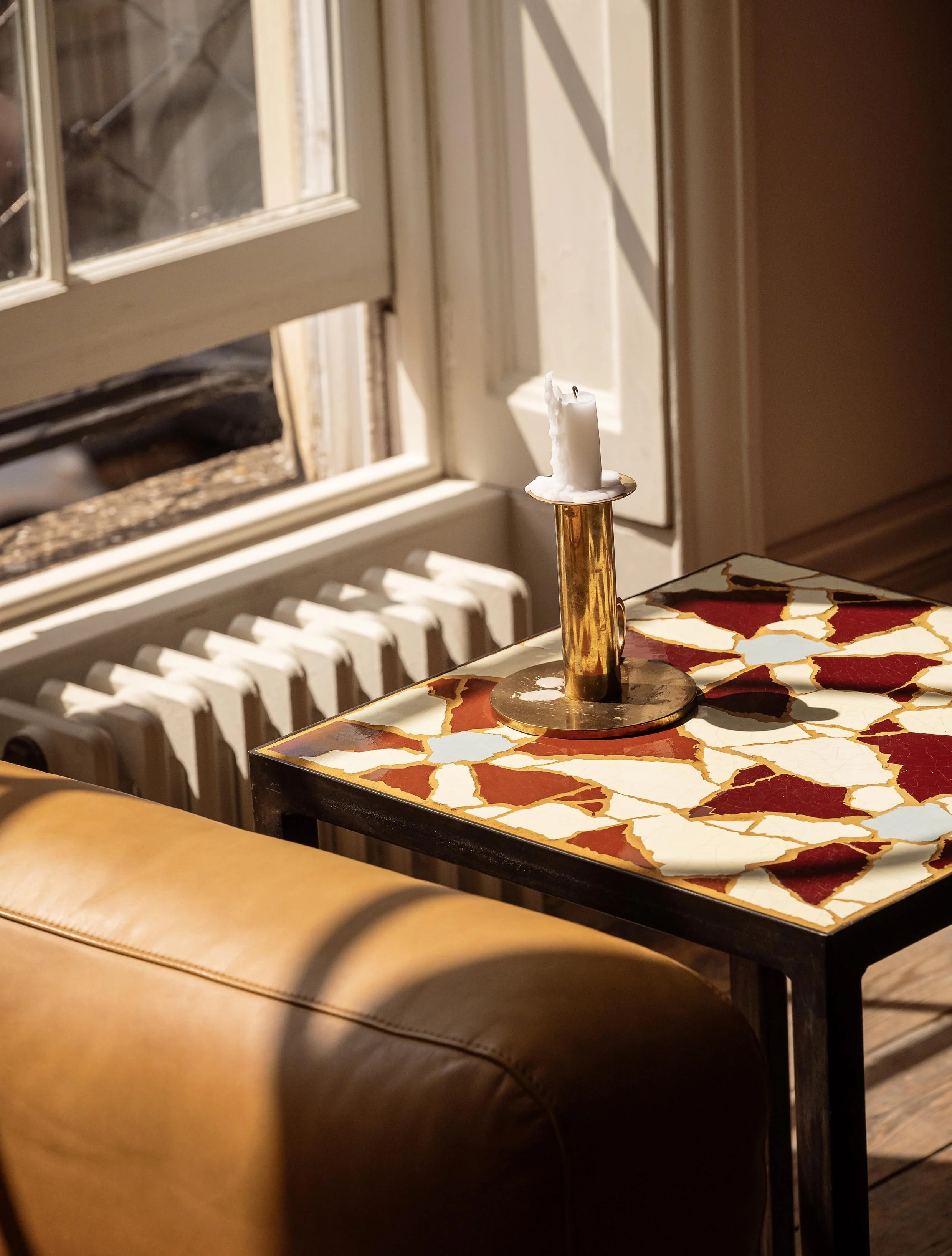 A table with a colorful, cracked tile surface featuring red, white, and orange hues, with a brass candle holder and a white candle on top. Part of a brown leather sofa is visible in the foreground, and a window with a radiator underneath is in the ba