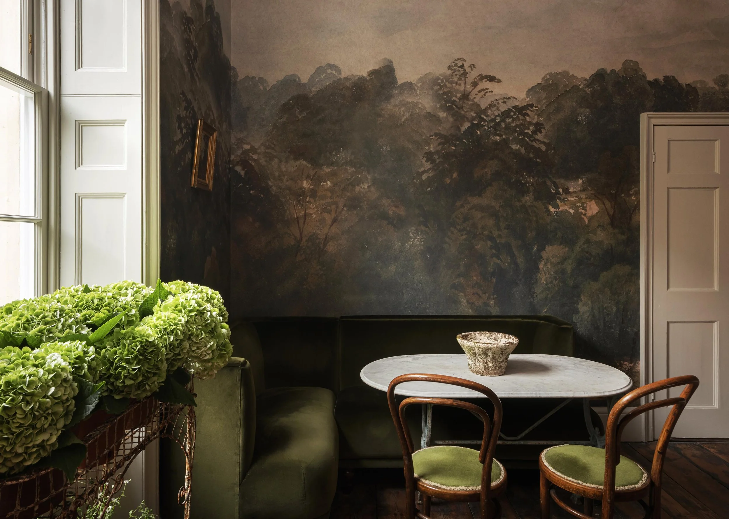Interior of a room with a green sofa, a round marble table, three wooden chairs with green cushions, a large window with white frames, a wall mural of a forest landscape, and a potted plant with green hydrangeas.