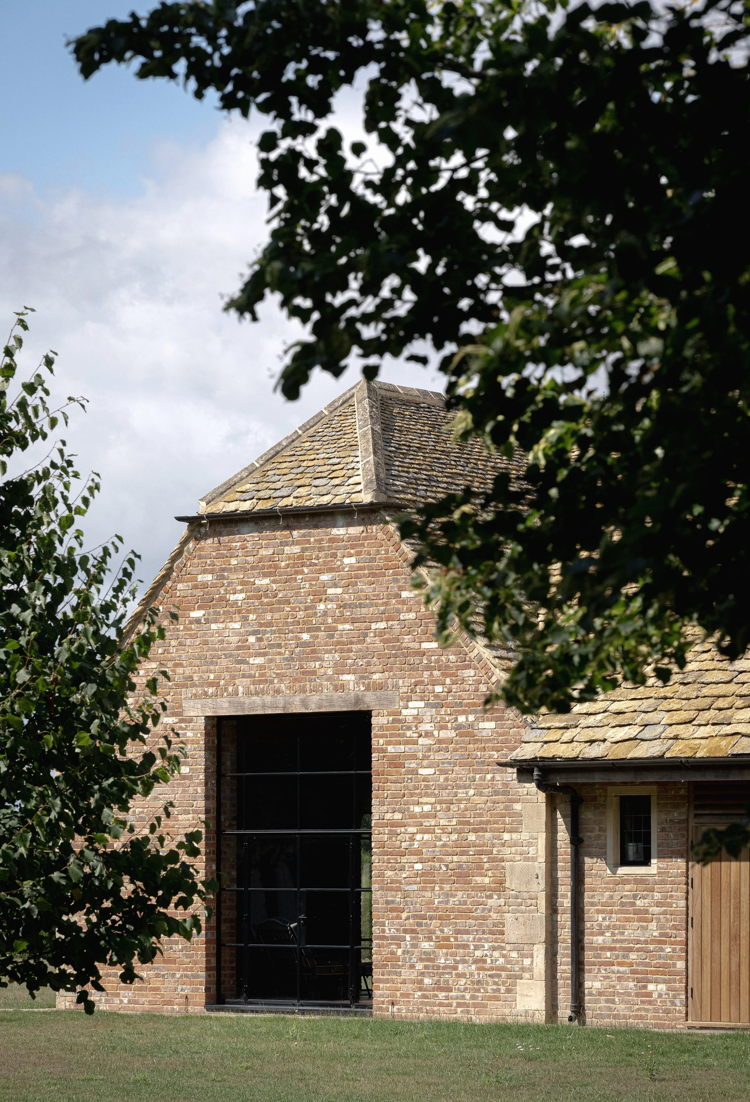 A brick building with a sloped tiled roof, partially obscured by leafy green trees, under a partly cloudy sky.