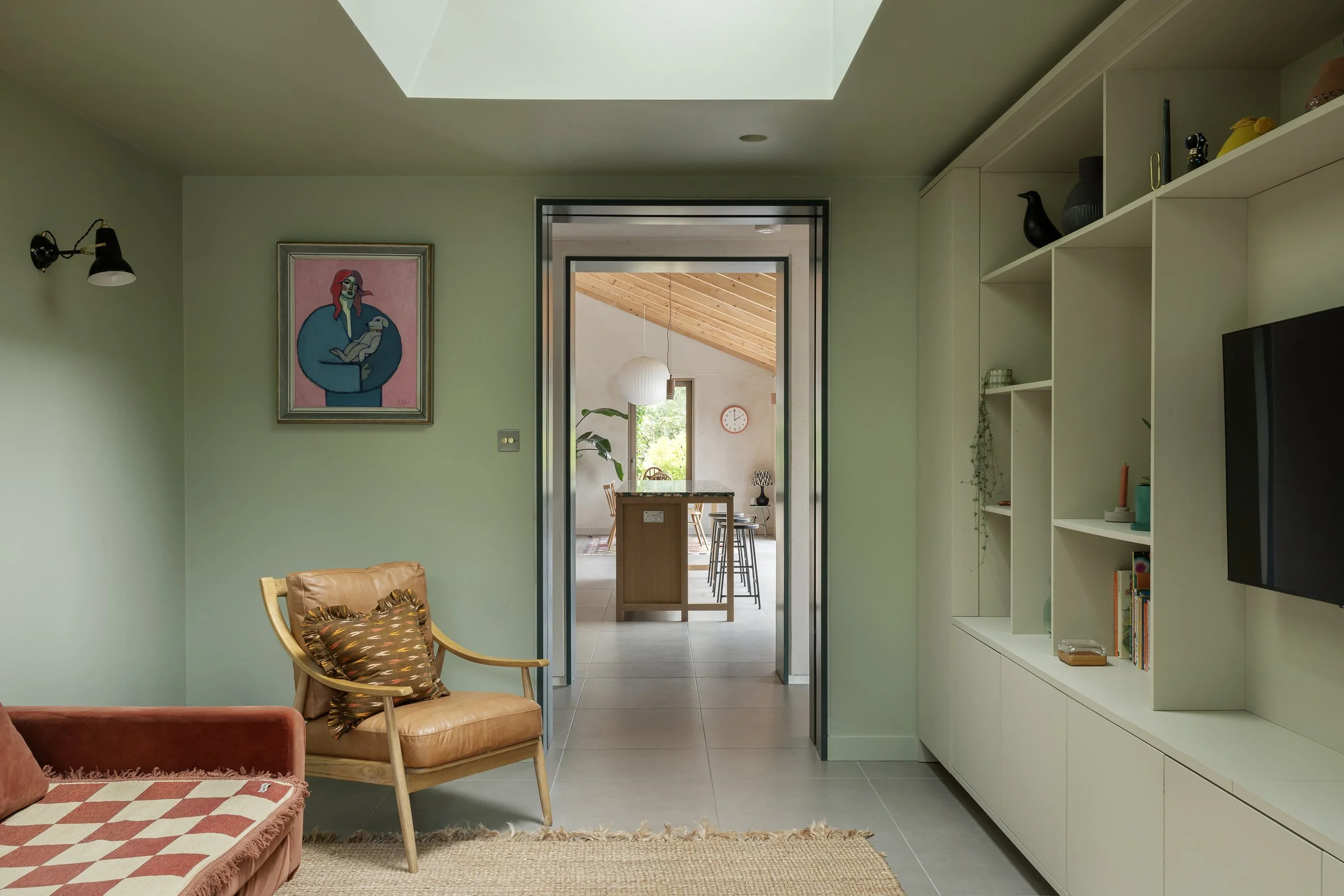 Living room with green walls, a framed artwork of a woman holding a bird, a tan armchair with a patterned pillow, built-in white shelves with decorative items, a wall-mounted TV, and a doorway leading to a kitchen area with a high ceiling and wooden 