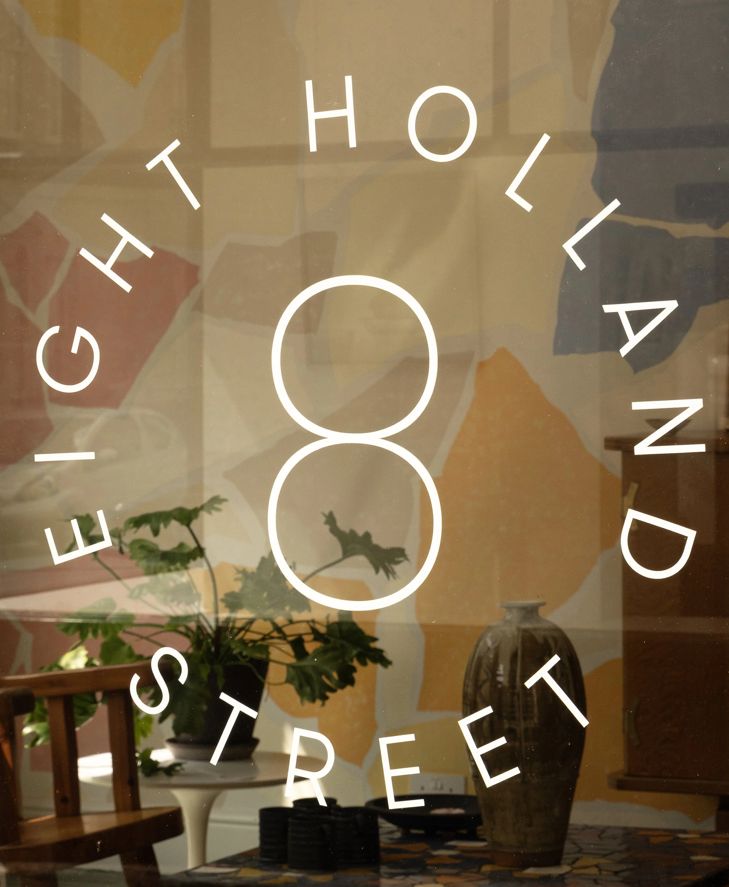 Reflective glass door with white text reading "EIGHTH HOLLANE STREET" and a large number 8 in the center. Behind the glass, indoor decor includes a potted plant, a wooden chair, a table with black plates, a vase, and a colorful abstract painting.