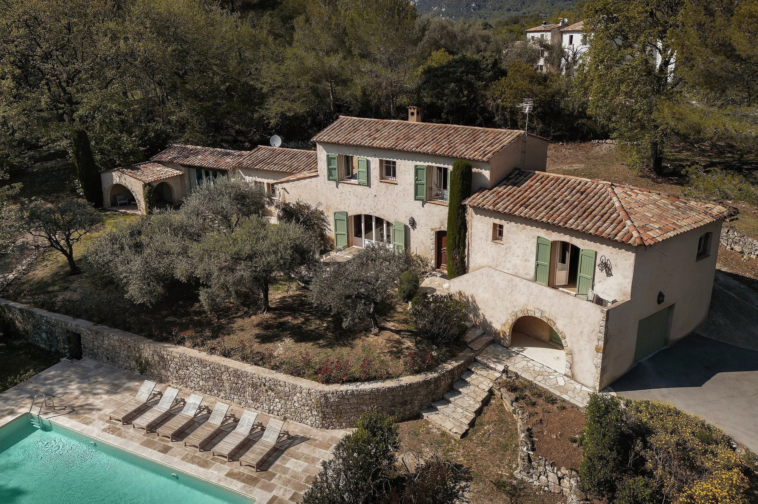 Aerial view of a Mediterranean-style house with a tiled roof, surrounded by trees, with a swimming pool with lounge chairs in the foreground.