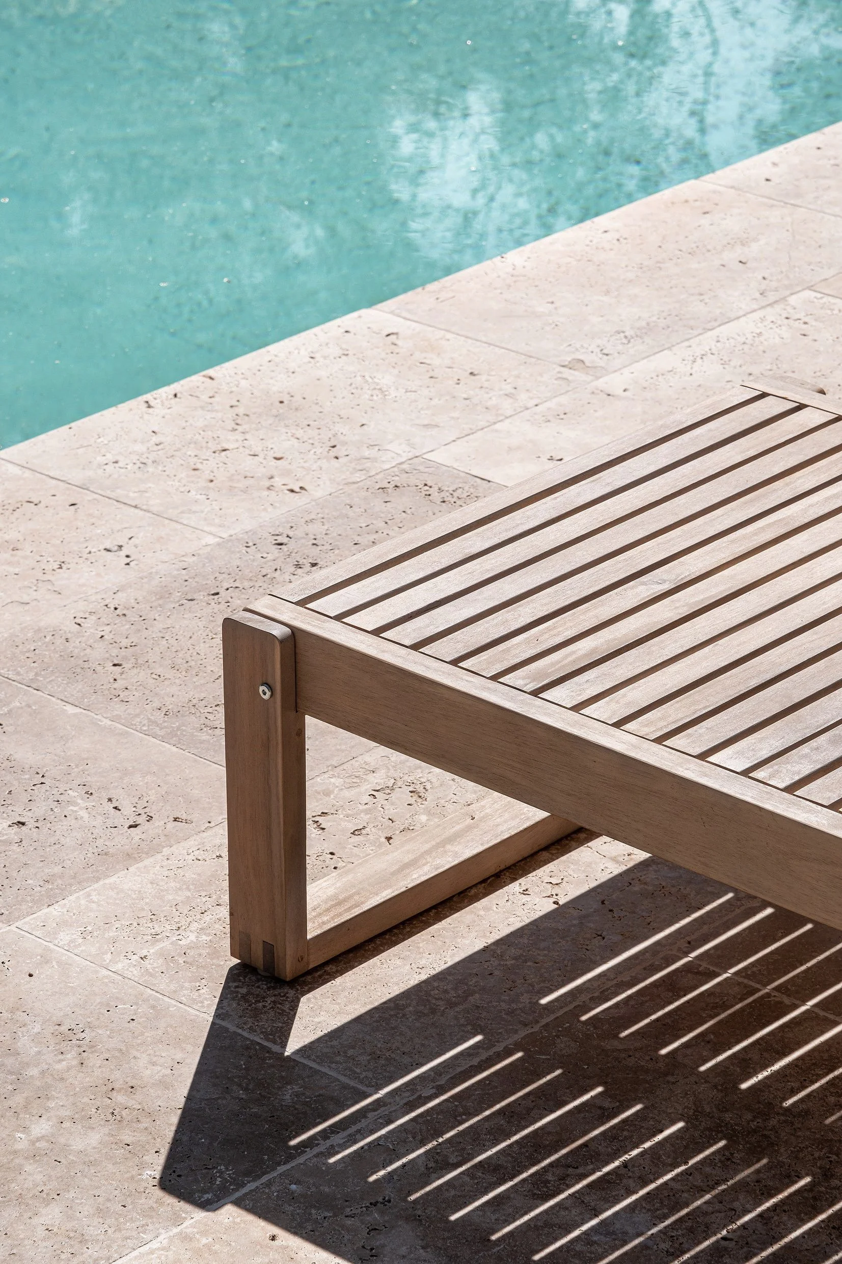 A wooden lounge chair by a swimming pool with sunlight creating striped shadows on the tiled pool deck.