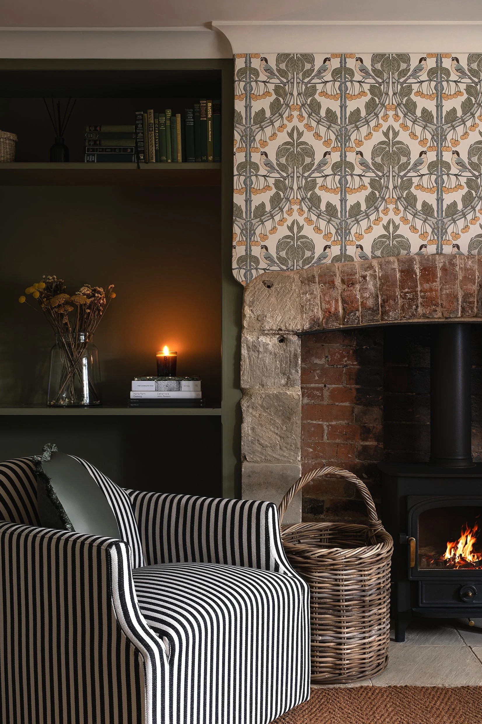 Cozy living room corner with a black stove, brick fireplace, striped armchair, wicker basket, bookshelf, candle, and patterned wallpaper.