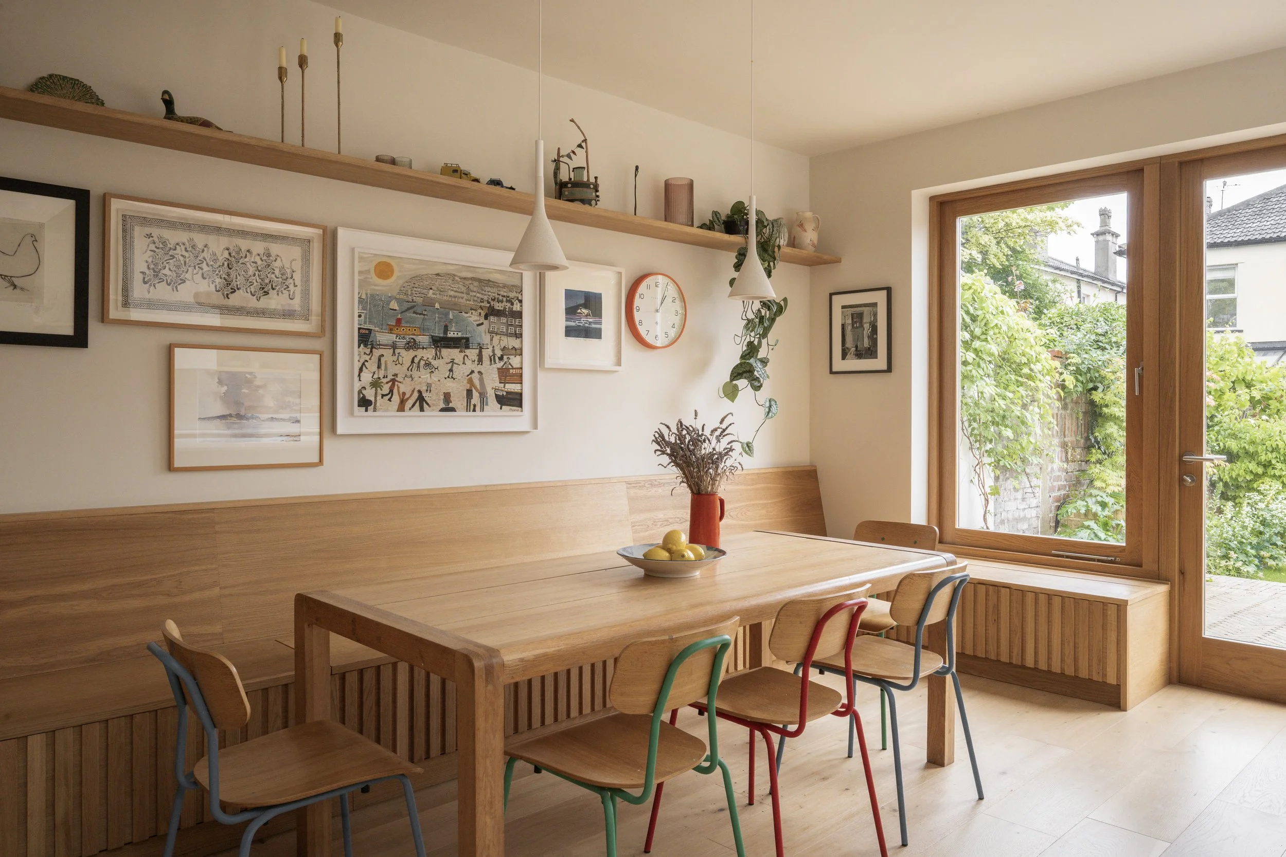 Wooden dining table with four colorful chairs, a vase with dried flowers, and a bowl of lemons. Bright room with large window, decorative wall art, hanging lamps, and a picture shelf.