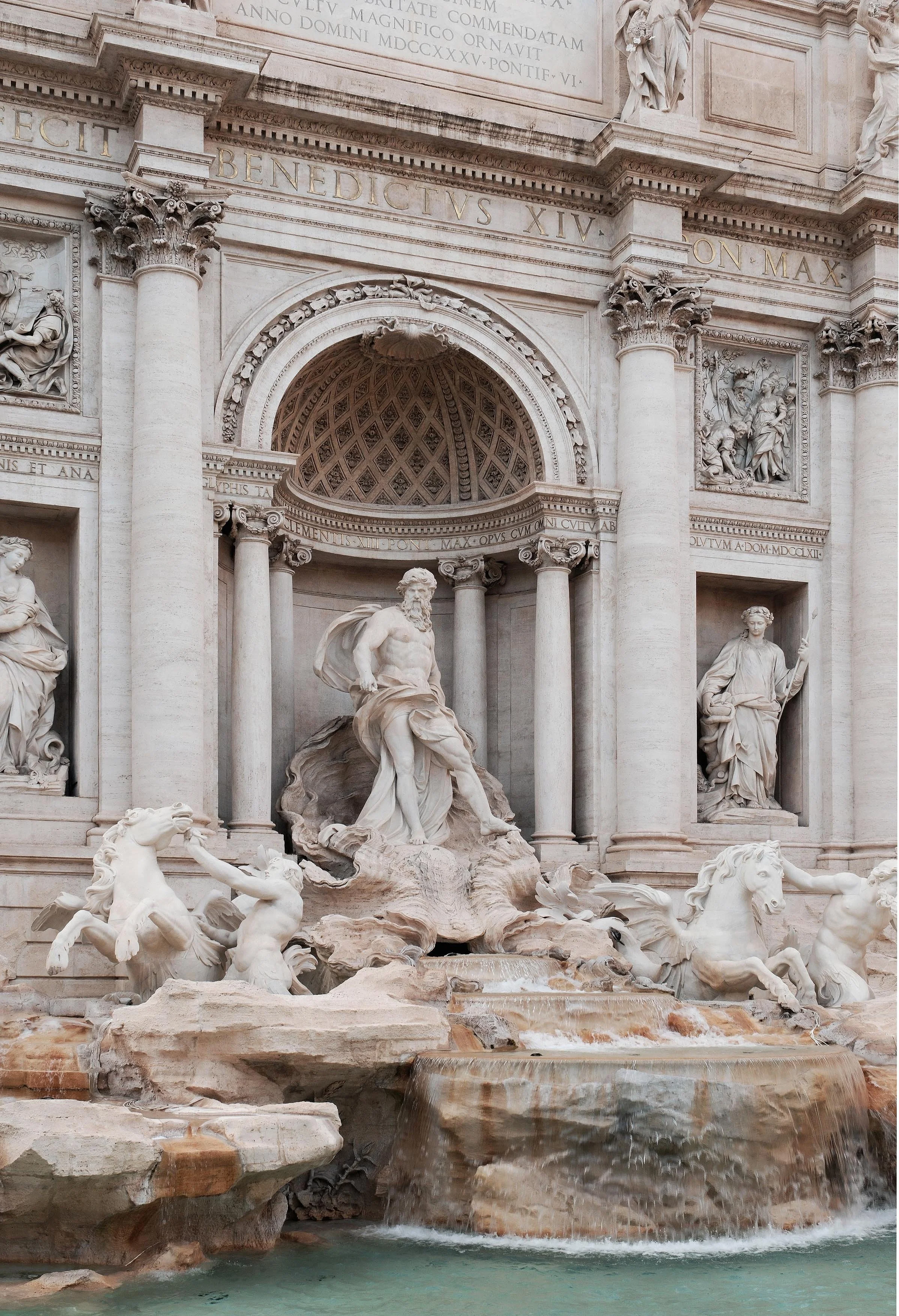 The Trevi Fountain in Rome, Italy, featuring a large central sculpture of Neptune with a flowing beard, surrounded by horses and mythological figures, with intricate architectural details and water flowing over rocks at the base.