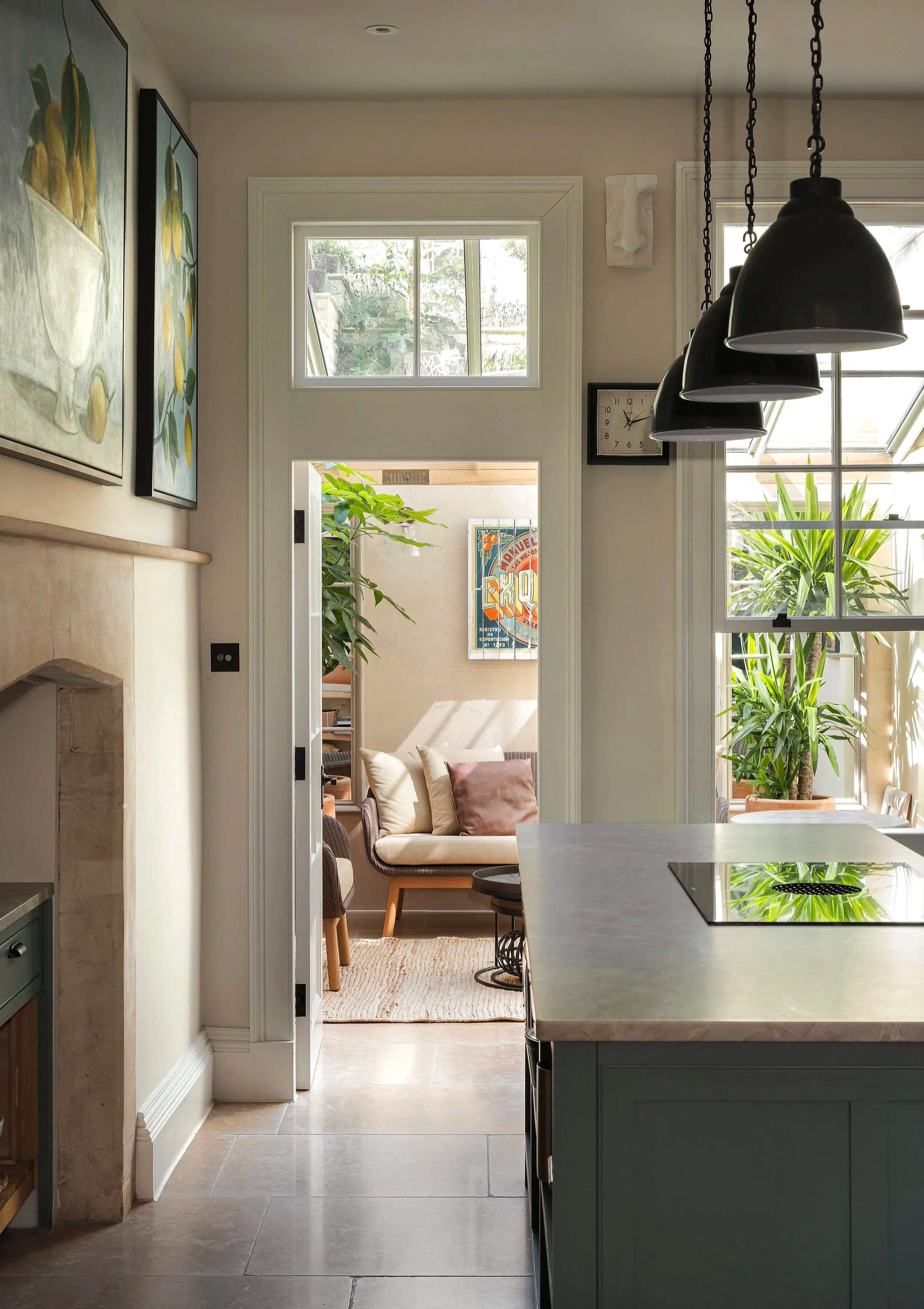 Interior view of a kitchen leading to a sunlit sitting room with a beige couch, large green plants, and framed artwork.