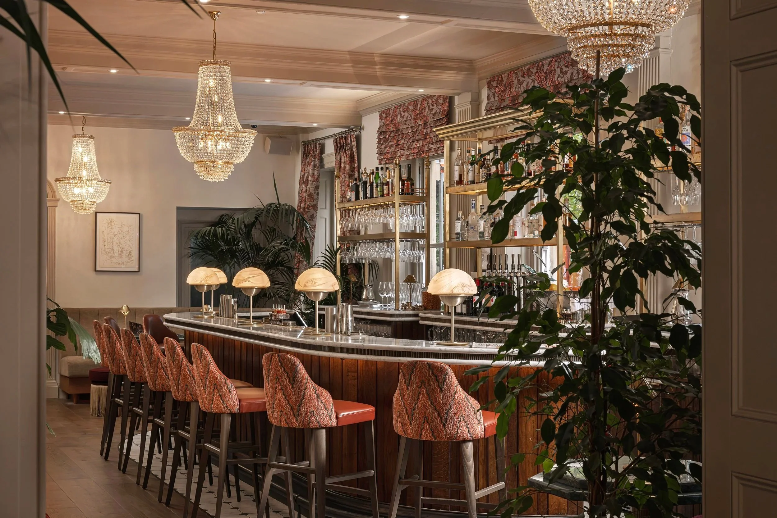 Elegant bar interior with chandeliers, a marble countertop, upholstered bar stools, and a well-stocked shelf of bottles and glassware, surrounded by lush green plants.