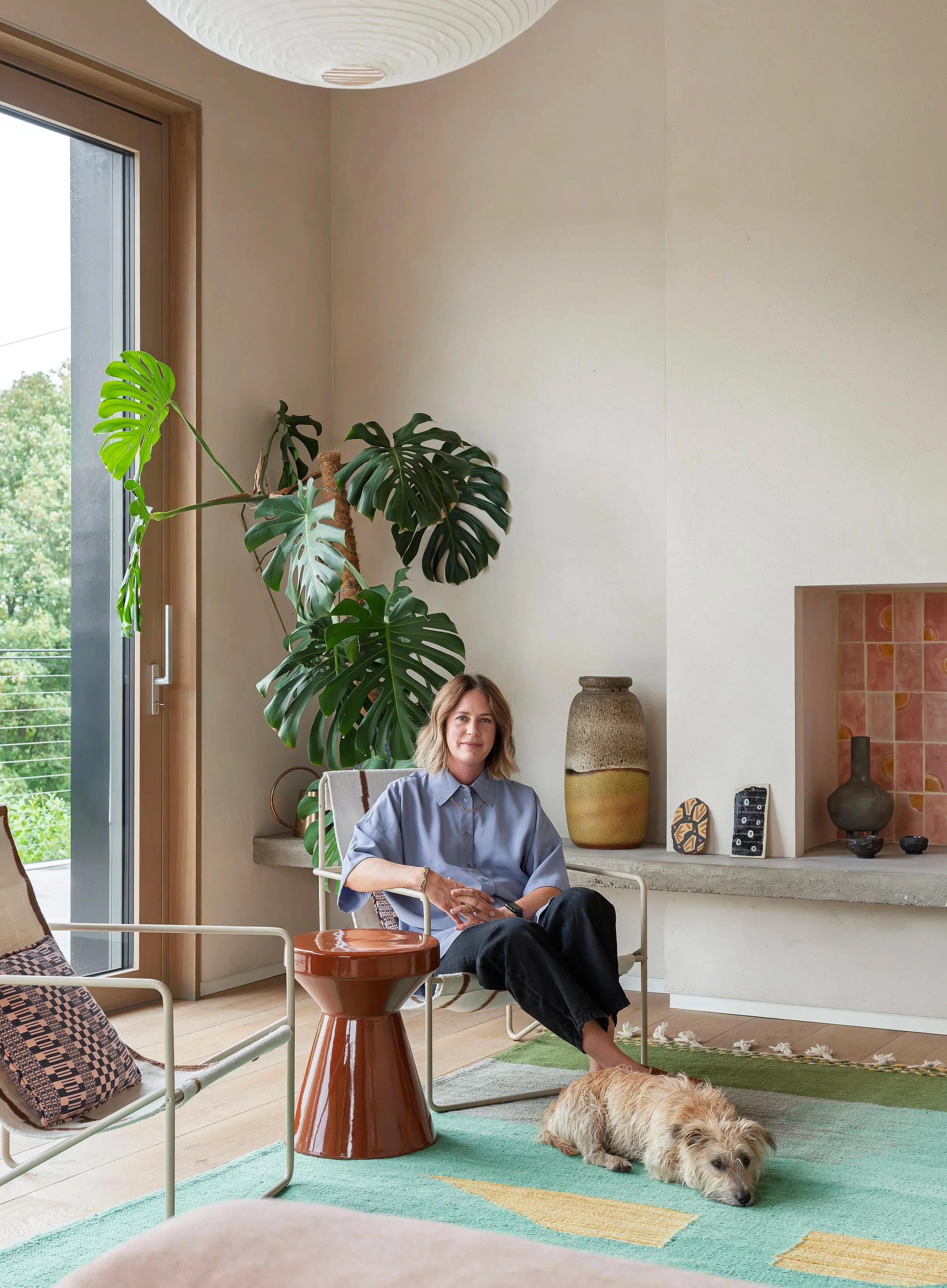 A woman sitting in a living room next to a dog on a green and yellow rug. There are large indoor plants, decorative vases, and artwork on the chimney breast. The room has a large window with a view of green trees outside.