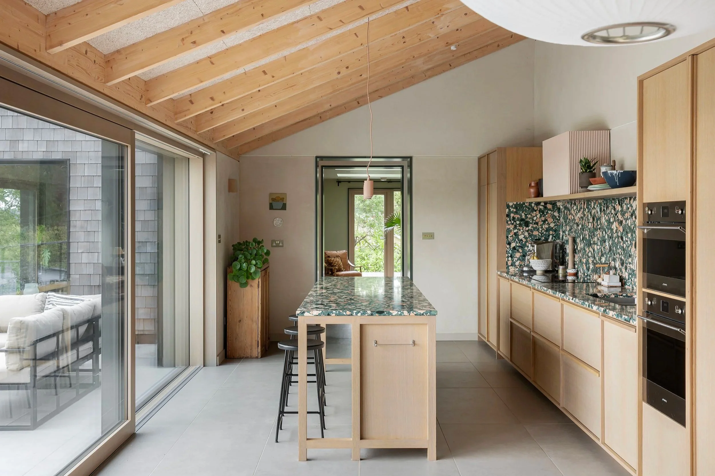 Modern kitchen with wooden cabinets, a marble island, and a patterned backsplash. Large sliding glass door leads to an outdoor patio with a sofa, and a view of trees outside. A doorway at the back shows a small seating area with a chair and a window