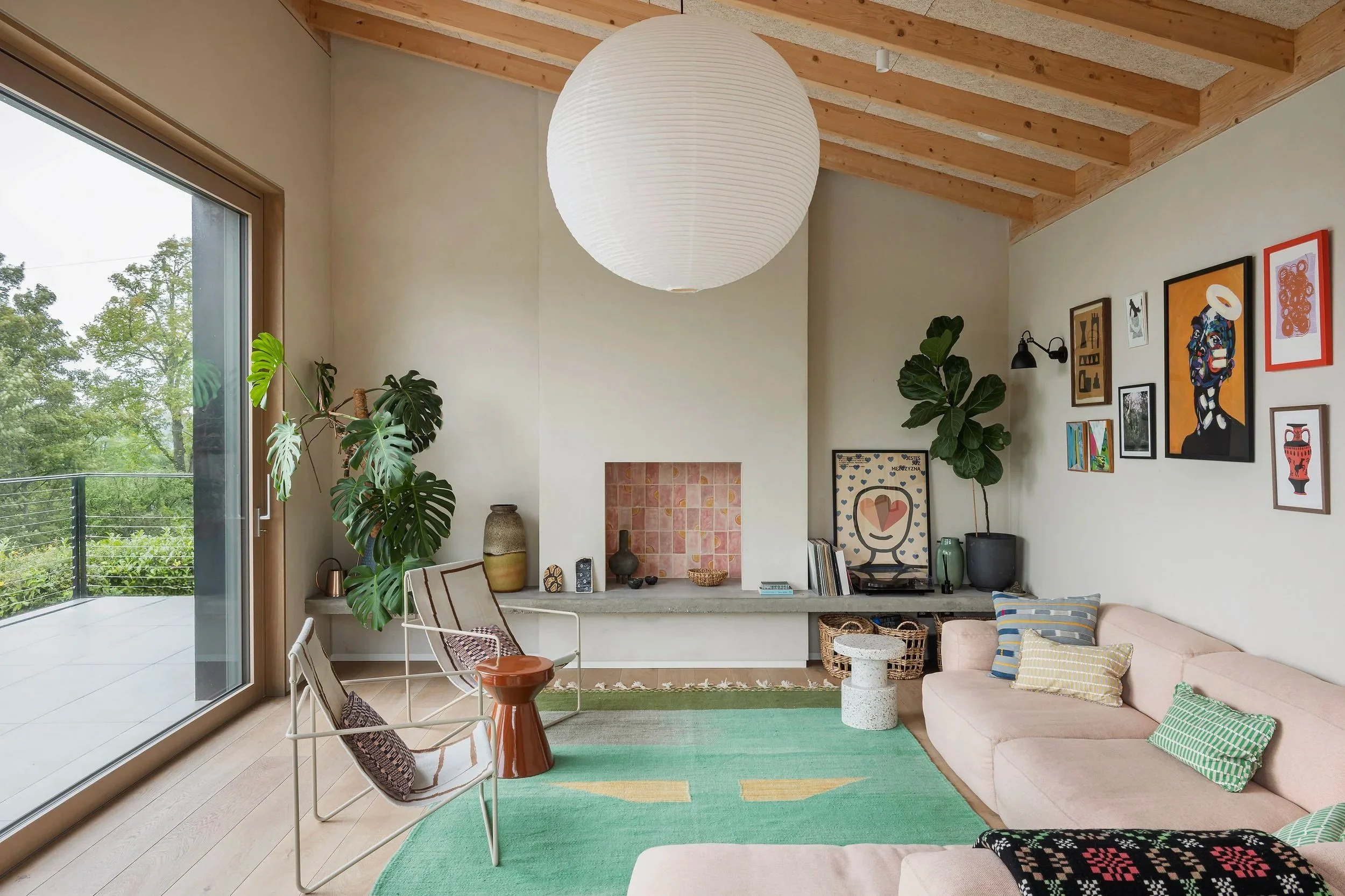 Living room with large hanging paper lantern, sliding glass door leading to balcony, green indoor plants, colorful artwork on the wall, pink sofa with striped pillows, green rug, and a low fireplace with decorative items.