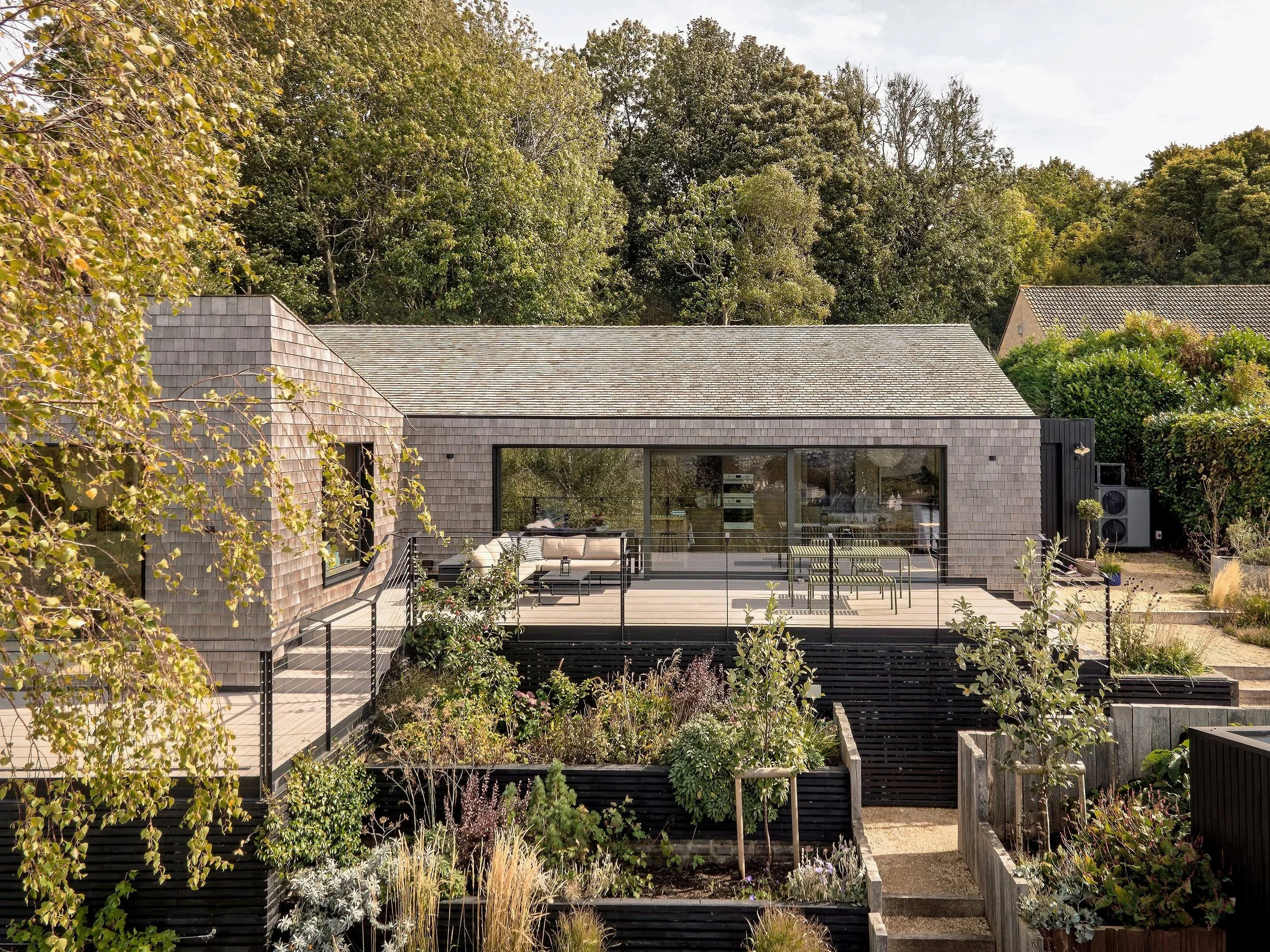 Modern house with a large wooden deck, glass sliding doors, surrounded by greenery and garden beds, with trees in the background.