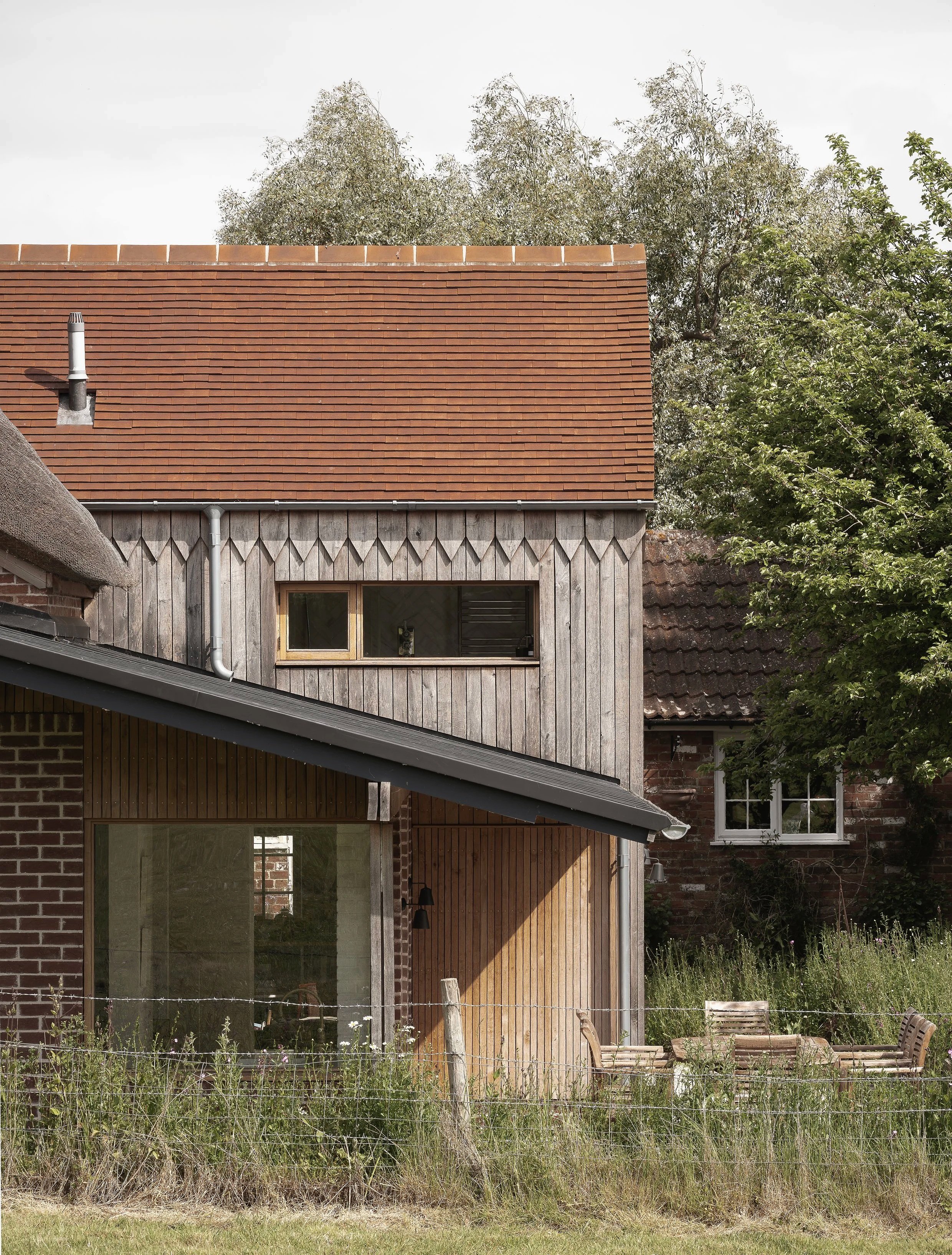 Residential house with a red tiled roof, wood and brick exterior, surrounded by greenery and outdoor seating.