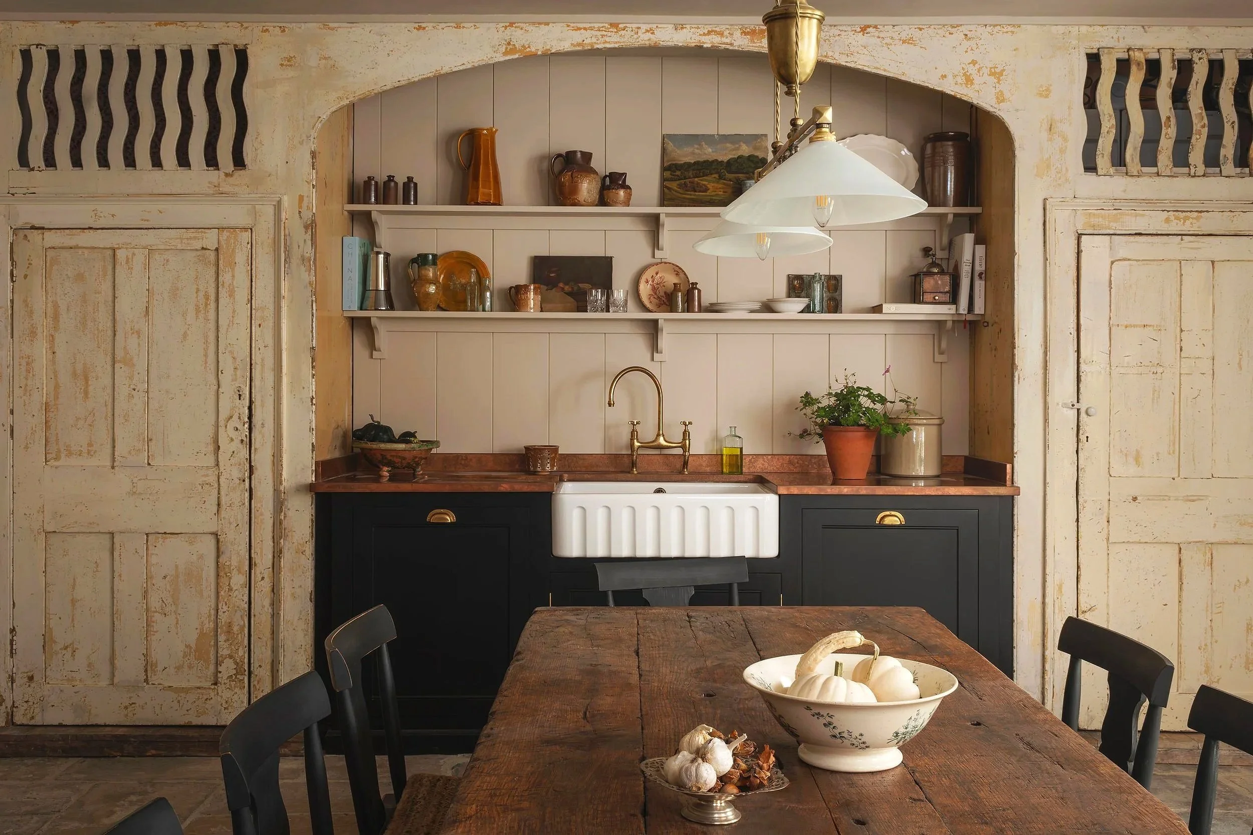 Rustic kitchen with a wooden dining table, black chairs, and open shelving with vintage pottery, books, and artwork. A farmhouse sink with a brass faucet is set into a red countertop, with a plant and kitchen essentials nearby. Vintage sliding doors 