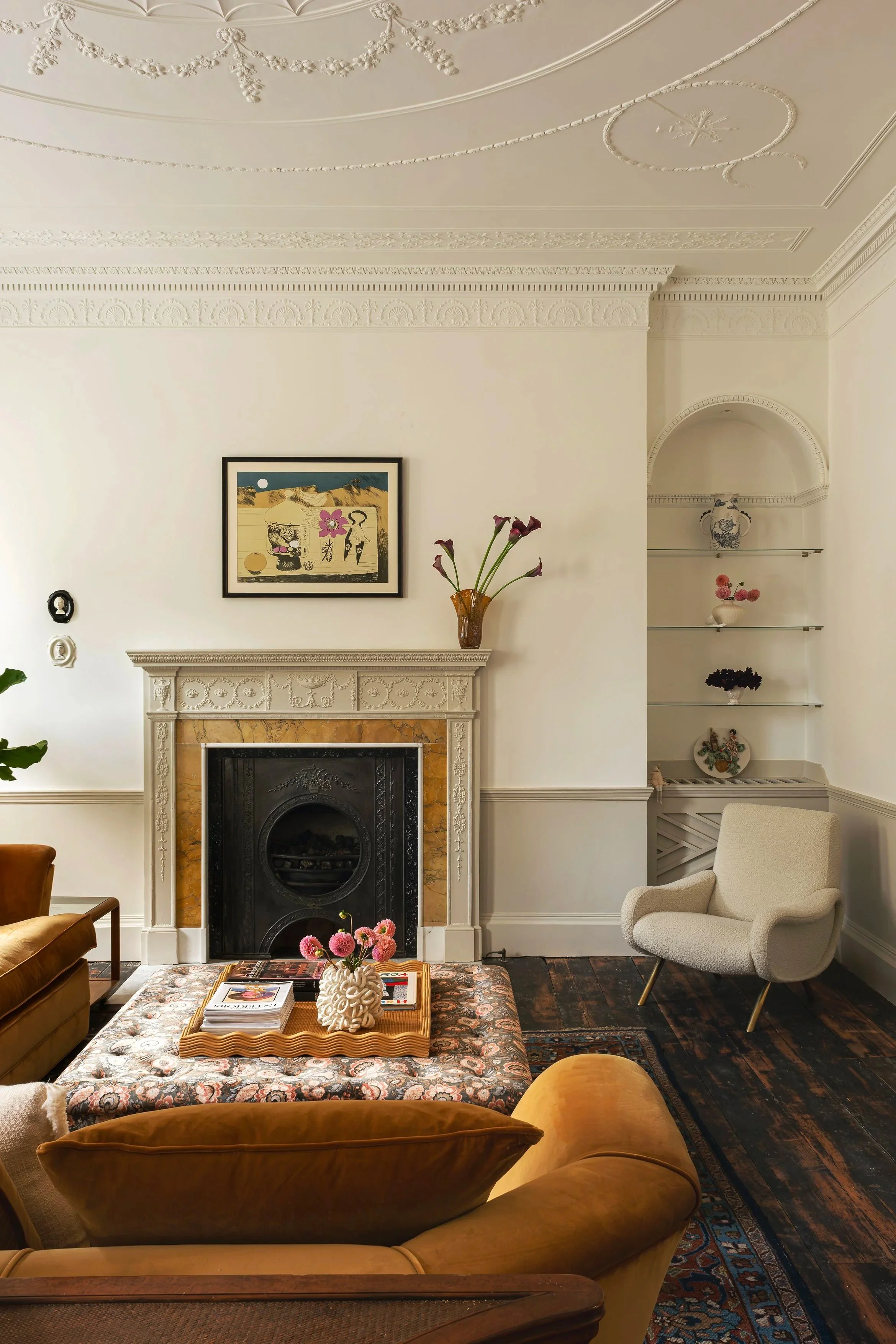 A cozy living room corner with a white ornate fireplace, a framed abstract artwork above, a yellowish sofa with a patterned ottoman holding books and a pink flower vase, a white armchair, and built-in shelves with decorative vases and flowers, all wi