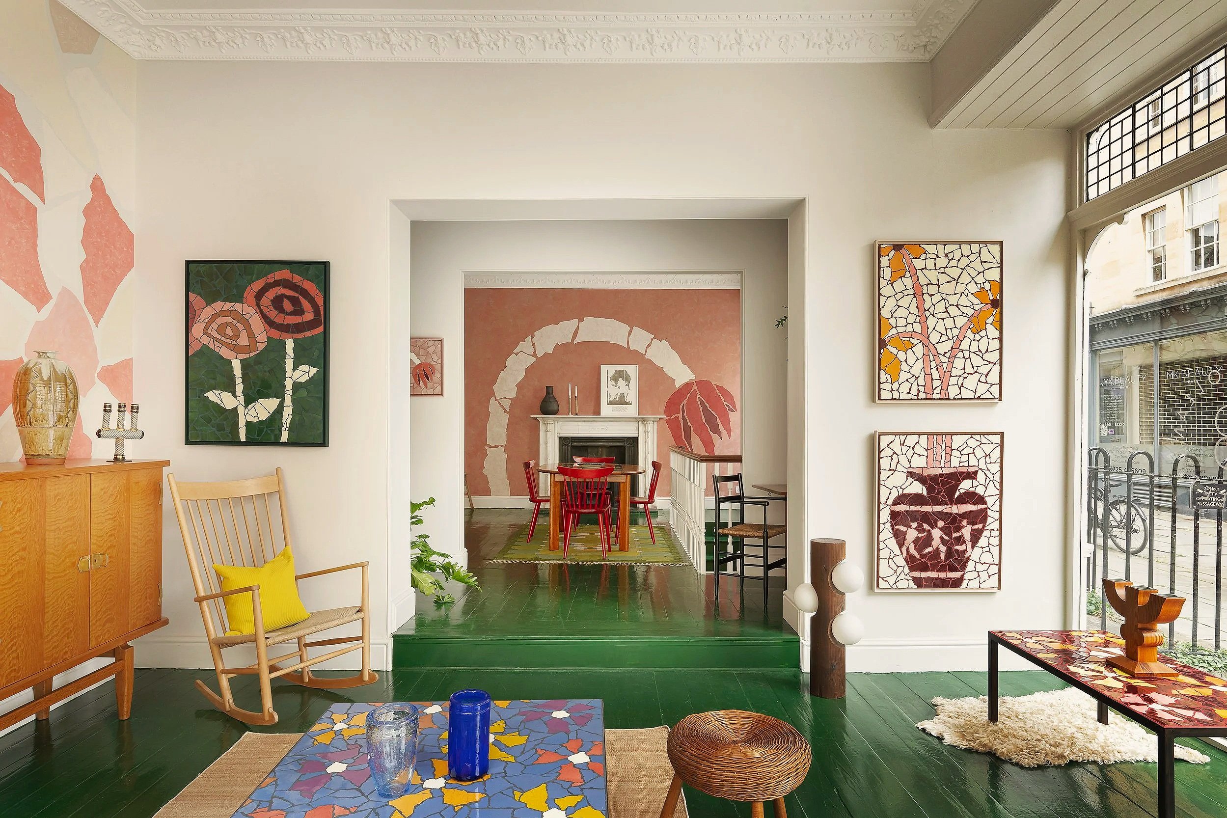 Interior of a home with colorful mosaic-style artwork on the walls, wooden furniture, and a view through large windows to the street.