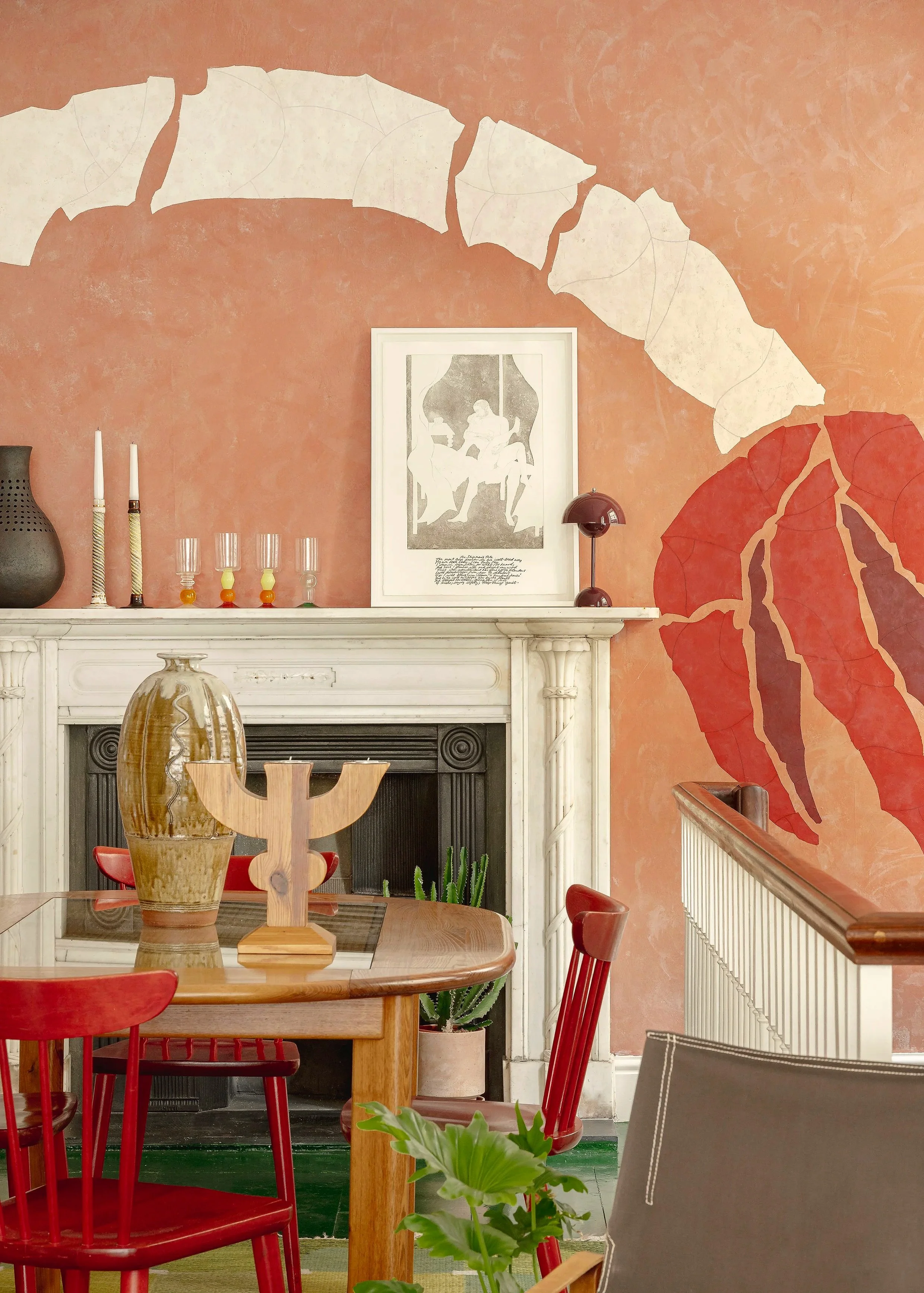 Interior of a room with a white fireplace mantel, an orange-pink wall with large leaf mural, a wooden dining table with red chairs, decorative objects on the mantel including vases, candles, and glasses, a framed art piece, and plants.