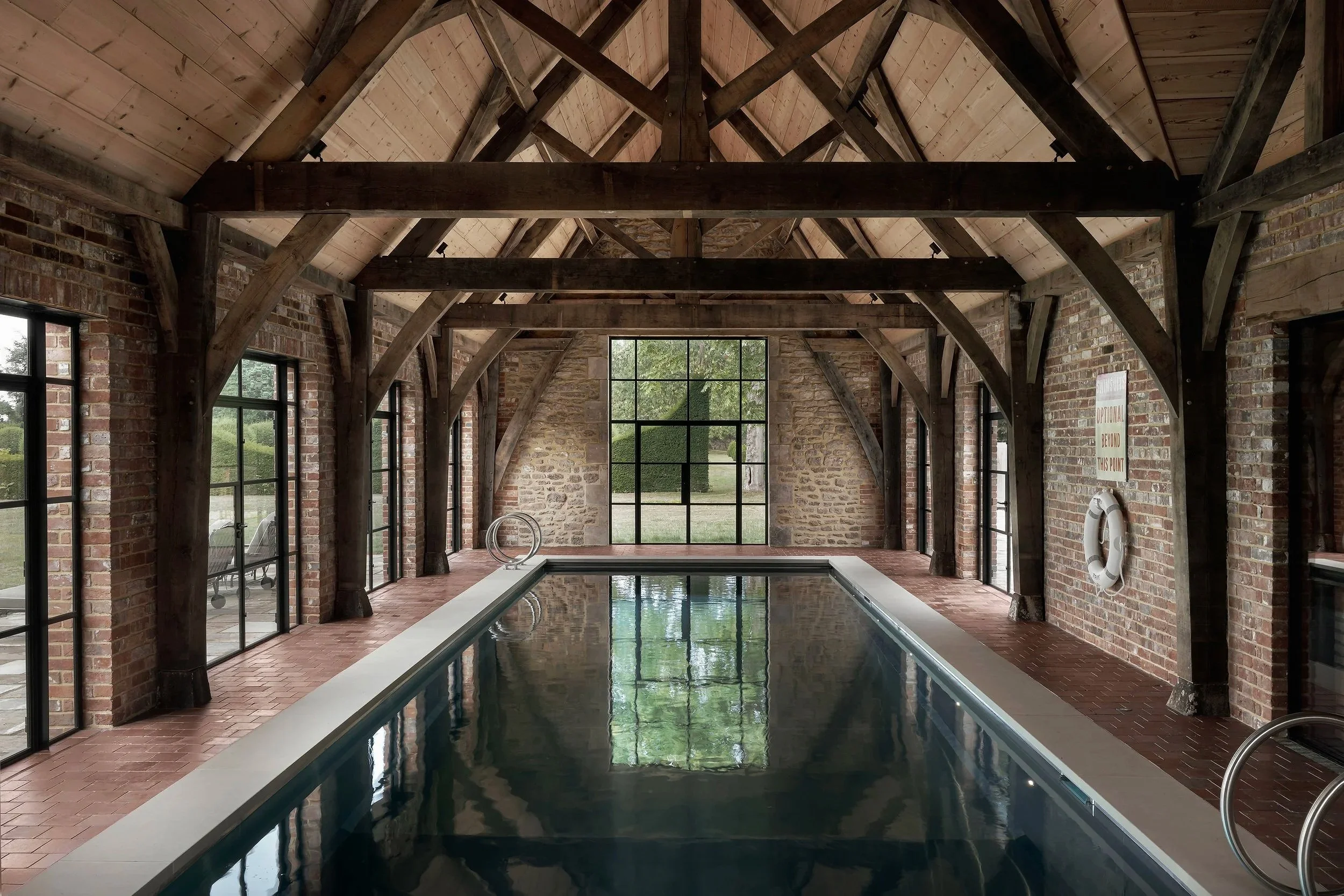Indoor pool area with brick walls and wooden beams, large window at the end reflecting the pool, sign on the wall, and life ring.