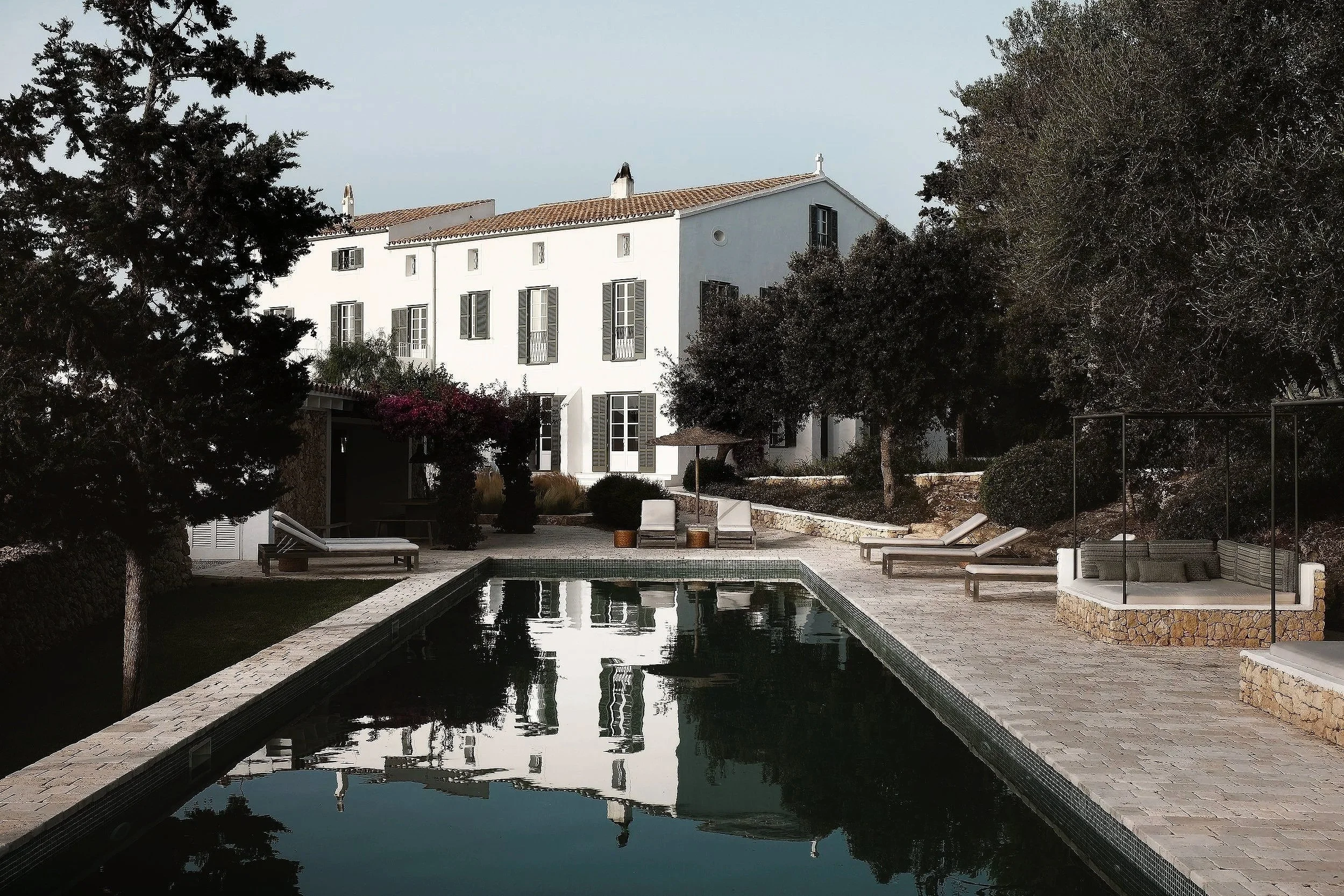A courtyard with a rectangular swimming pool, surrounded by lounge chairs and greenery, with a large white building in the background.