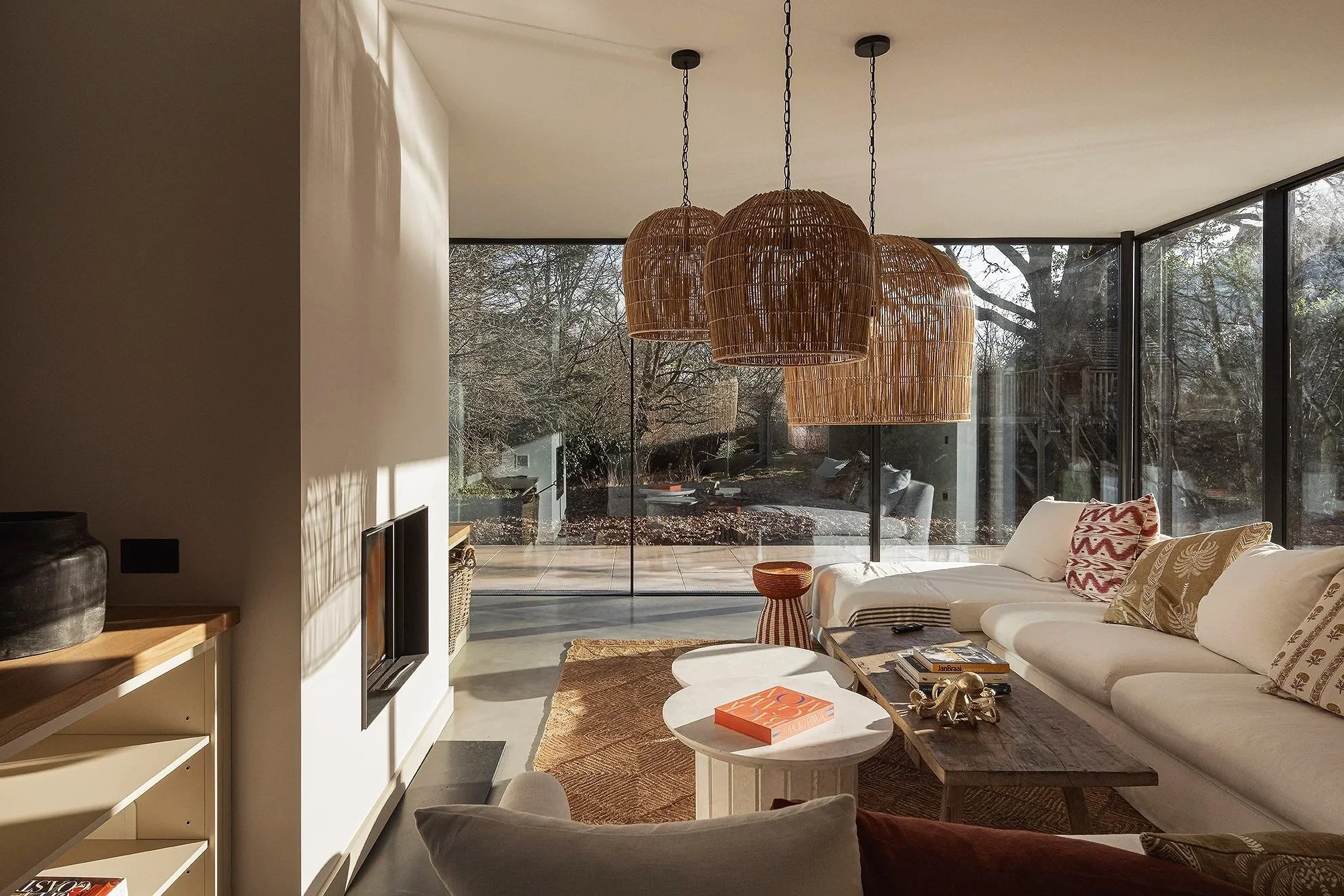 Living room with large glass windows, wooden pendant lights, white sectional sofa with patterned pillows, coffee table with books, small side table, and a view of outdoors with trees.