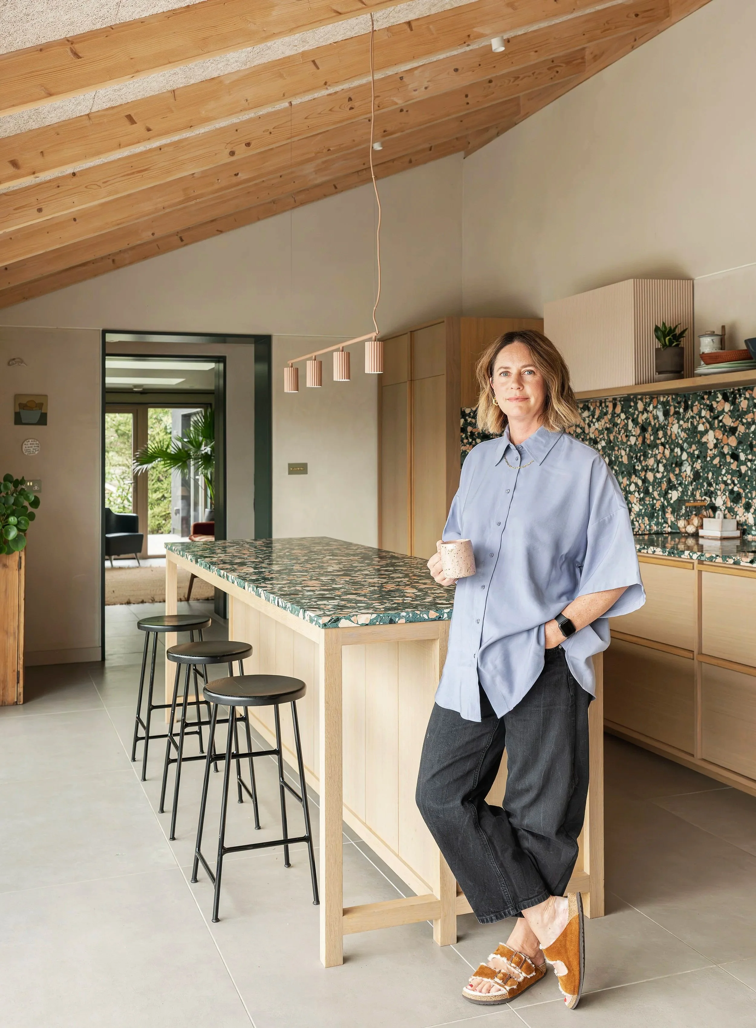 A woman standing in a modern kitchen, holding a mug, wearing a light blue oversized shirt, black jeans, and slippers. The kitchen has a marble island, wooden cabinets, and a sloped wooden ceiling.