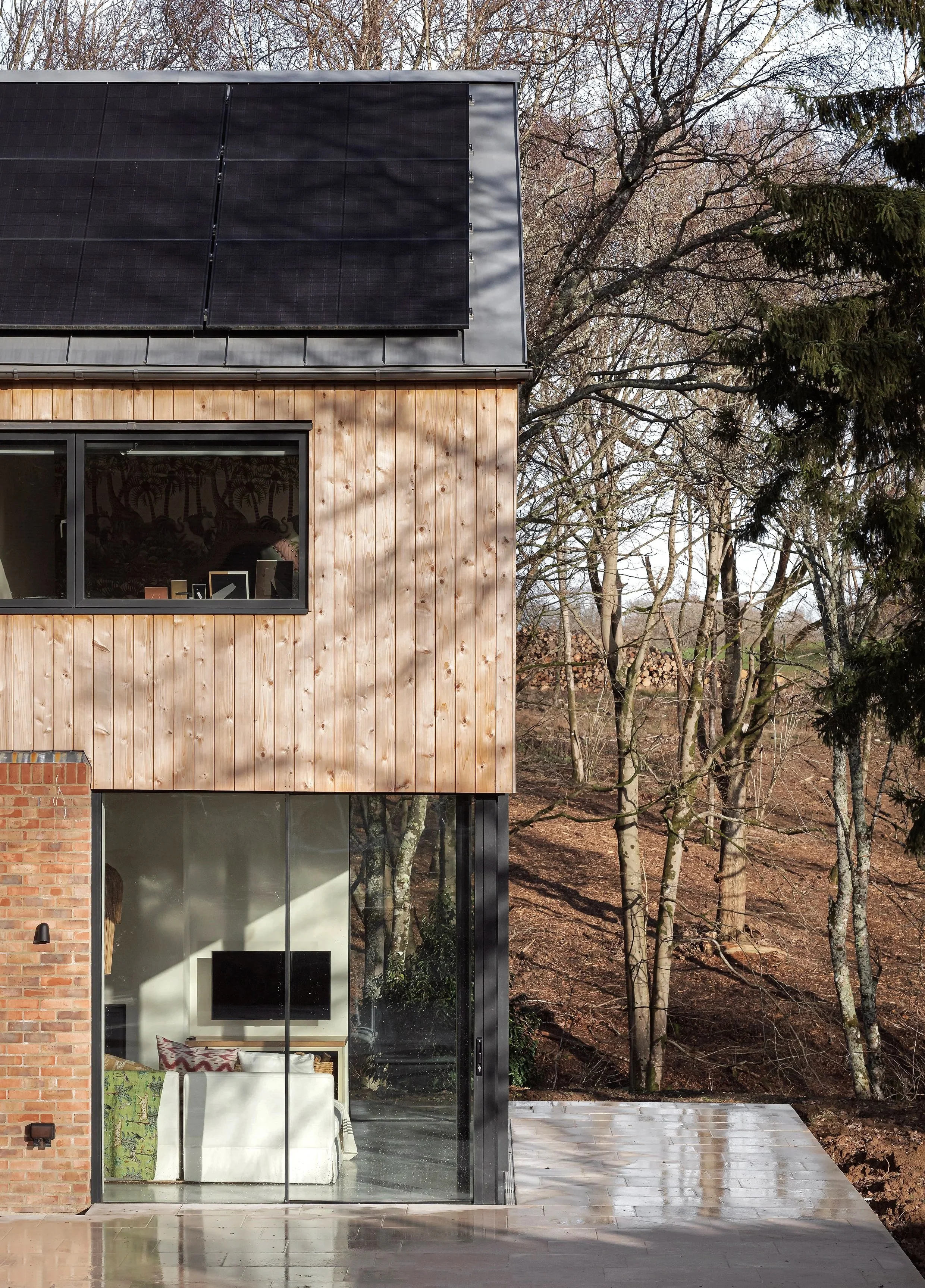 Modern house with wood-paneled upper exterior, brick lower exterior, and large glass window revealing a living room with a TV and sofa inside. The roof has black solar panels, and there are leafless trees in the background.