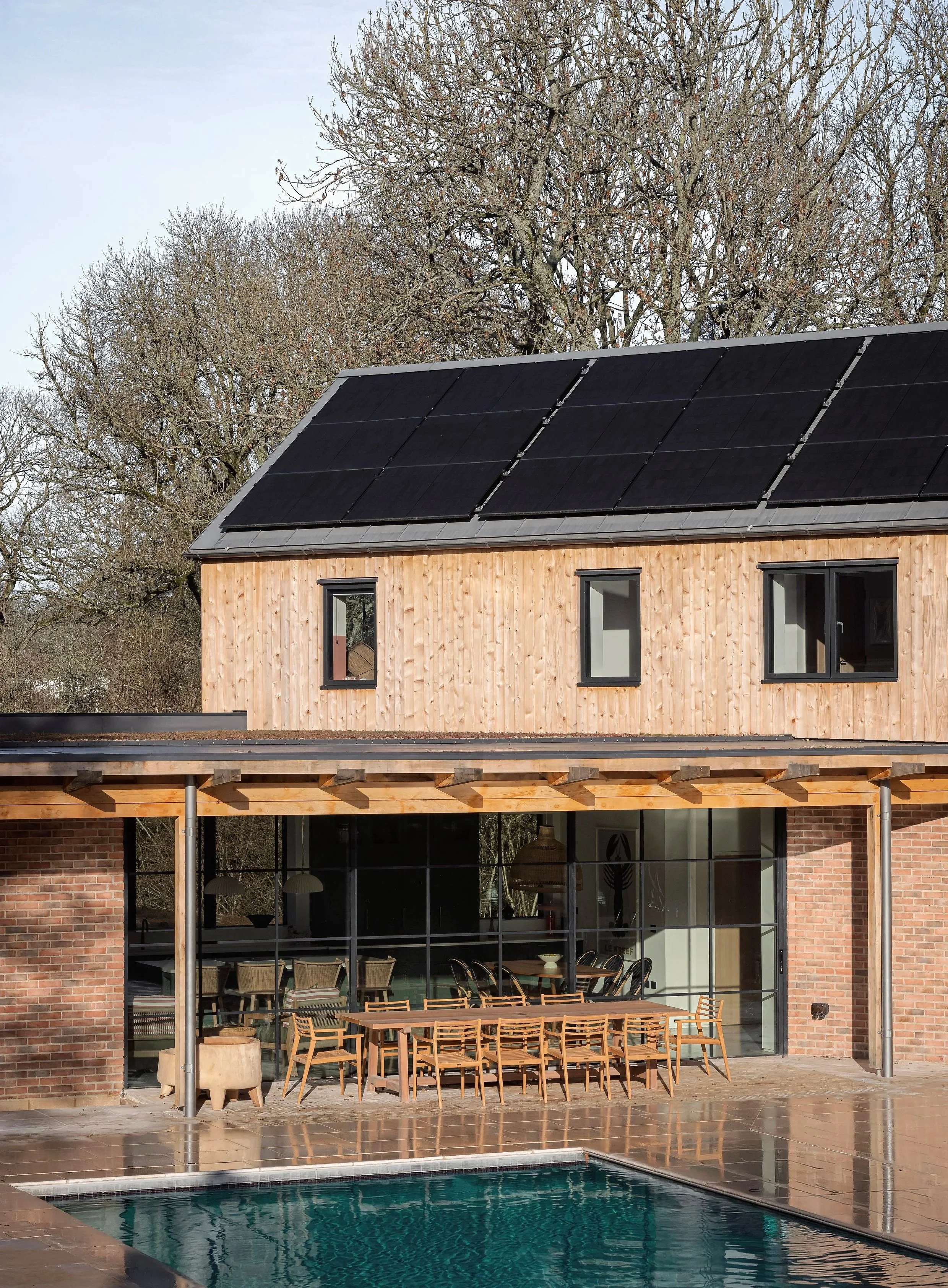 Modern house with solar panels on the roof, outdoor dining area, pool, and large glass sliding doors with trees in the background.