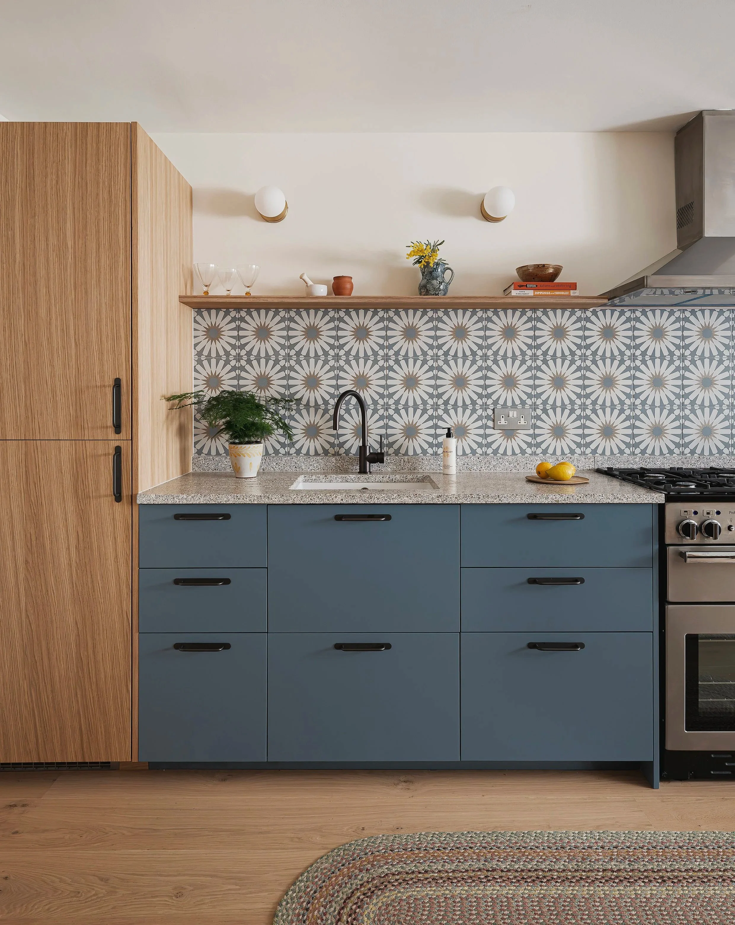 Kitchen with blue cabinets, a granite countertop, and a patterned backsplash. There is a black faucet, a potted plant, and a bottle of soap near the sink. The wall has two white round lights, open shelving with decorative items, and a stove on the ri