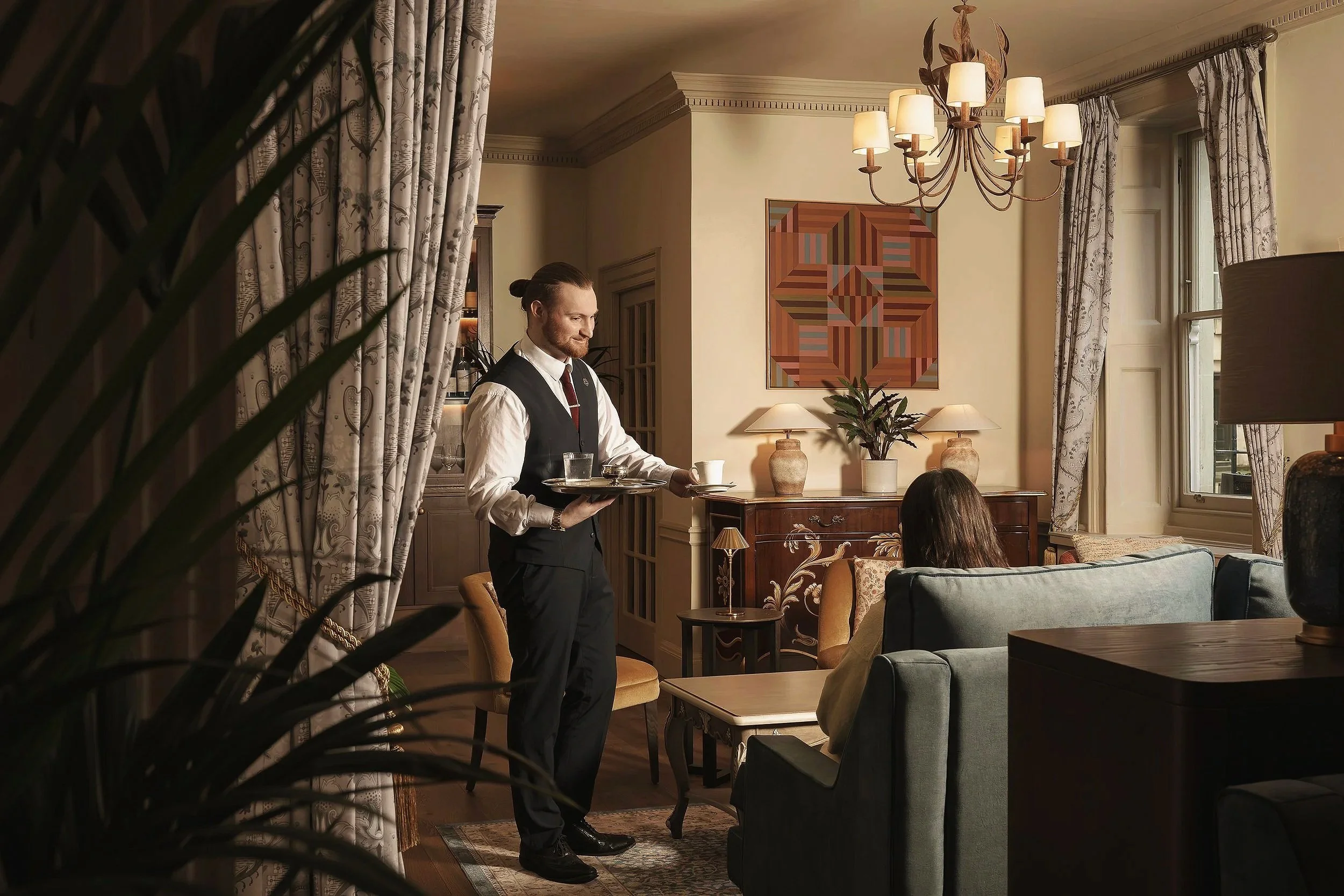A waiter in a black vest and white shirt serving drinks in a cozy, well-decorated living room with beige walls, curtains, framed artwork, and a chandelier.