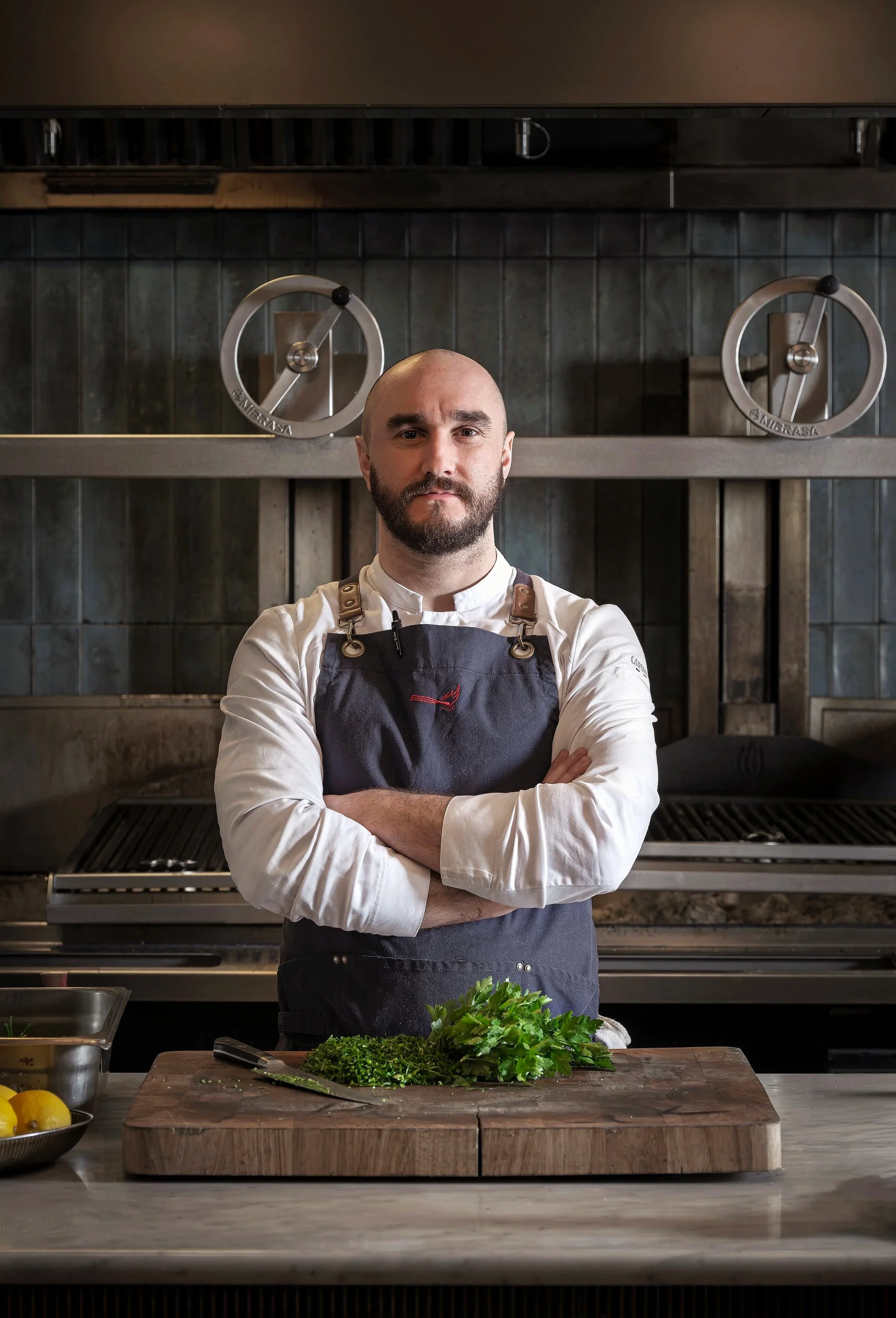 A male chef with a beard standing with arms crossed in a professional kitchen, with a chopping board and herbs in front of him.